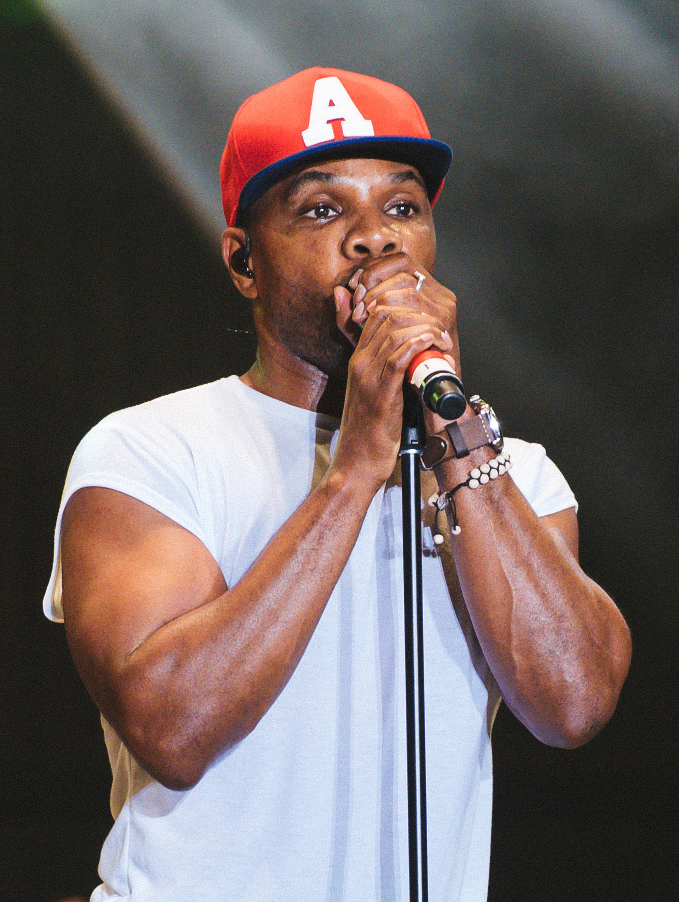 Kirk Franklin sings into a microphone on a stand with both hands around it, in a red baseball cap with a white A and a white T-shirt.