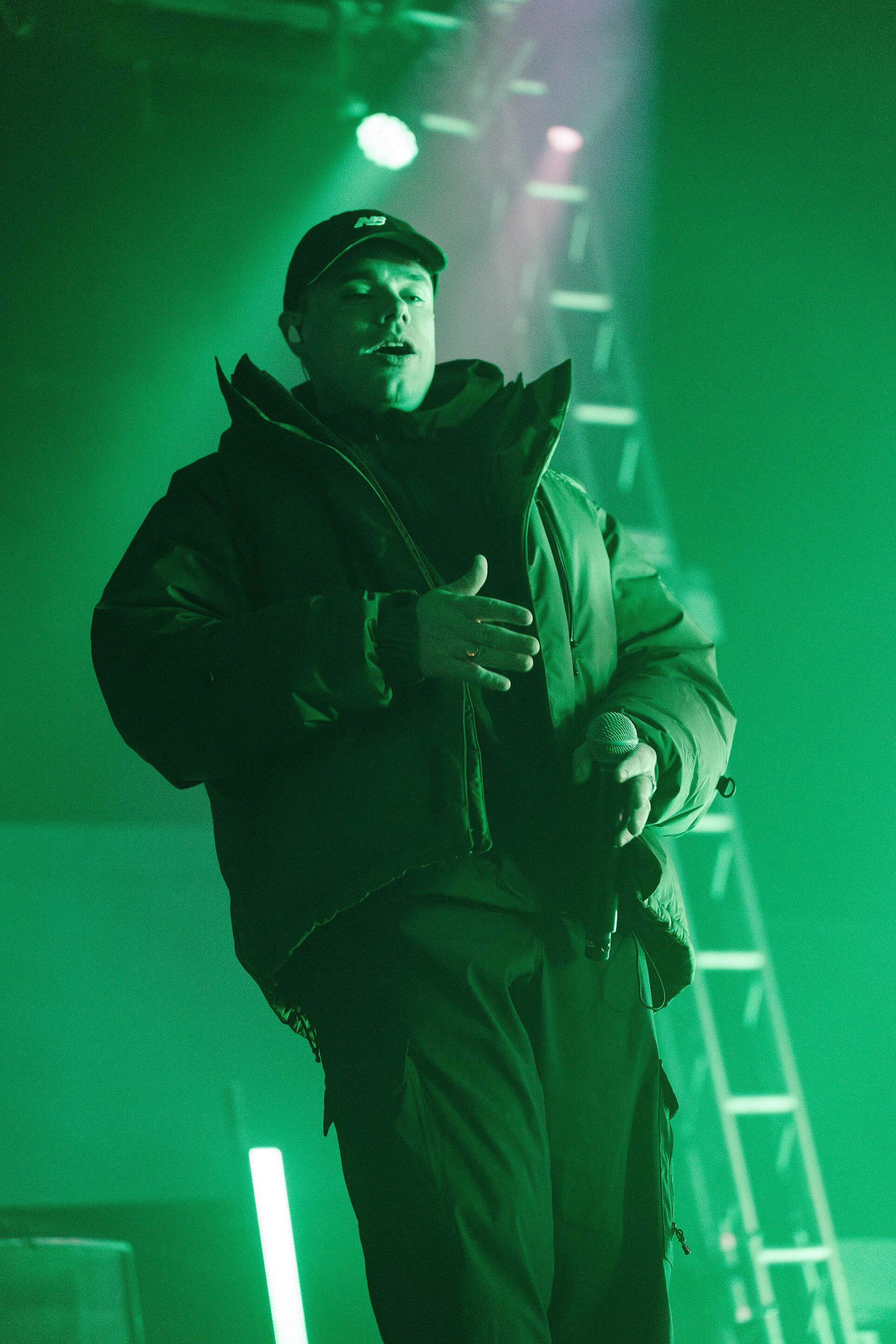 A Kneecap rapper in a padded jacket and cap holds a microphone low, singing under heavy green stage light.