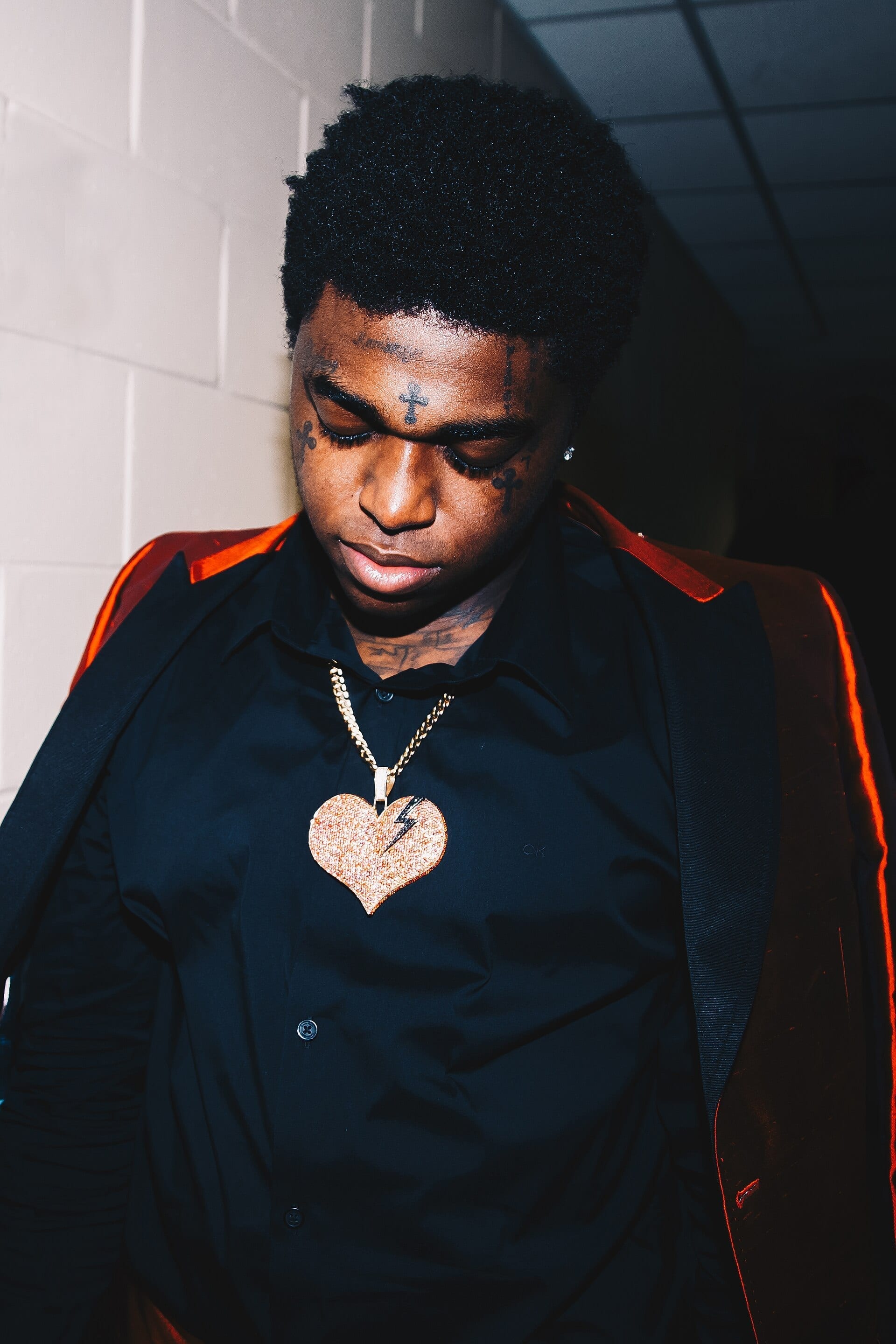 Kodak Black looks downward in a dim hallway, his face and neck marked with several tattoos, wearing a black button-up shirt under a jacket with red-lined lapels and a heart-shaped pendant necklace.