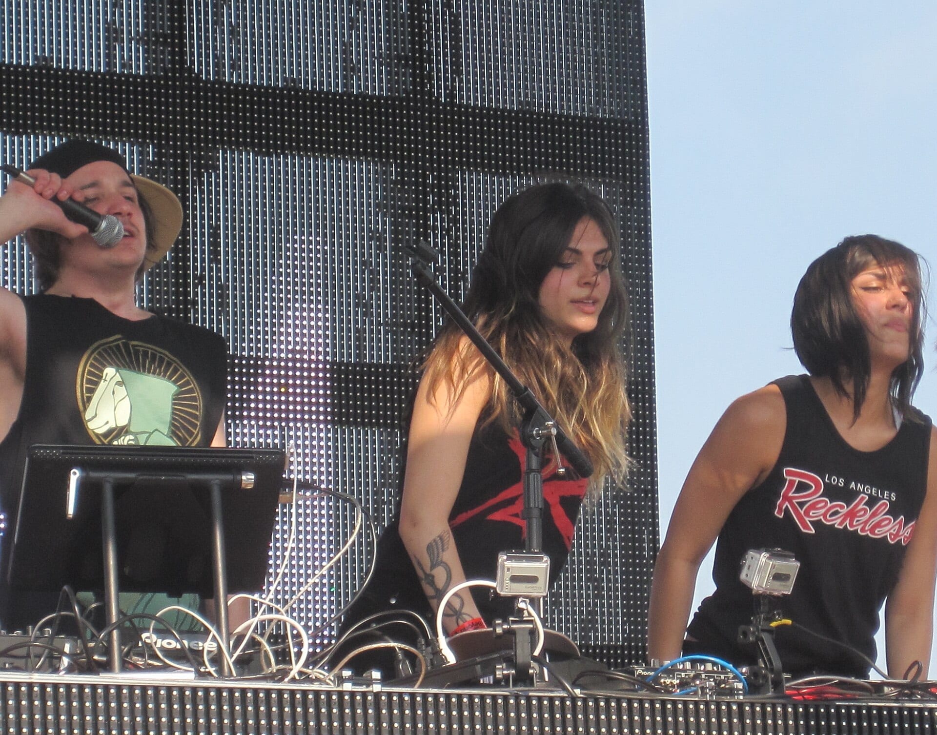 The three members of Krewella perform behind a DJ booth outdoors: a man in a wide-brimmed hat holds a microphone on the left, with Jahan Yousaf and Yasmine Yousaf beside him.