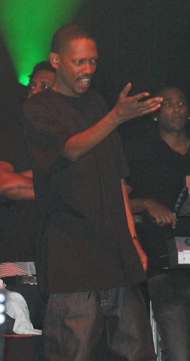 Kurupt gestures with both hands while speaking under green stage lighting, wearing a dark T-shirt, with other people visible behind him.