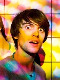 A close-up of Sam Eastgate, wide-eyed and open-mouthed in a candid moment, lit by colorful stage lighting.