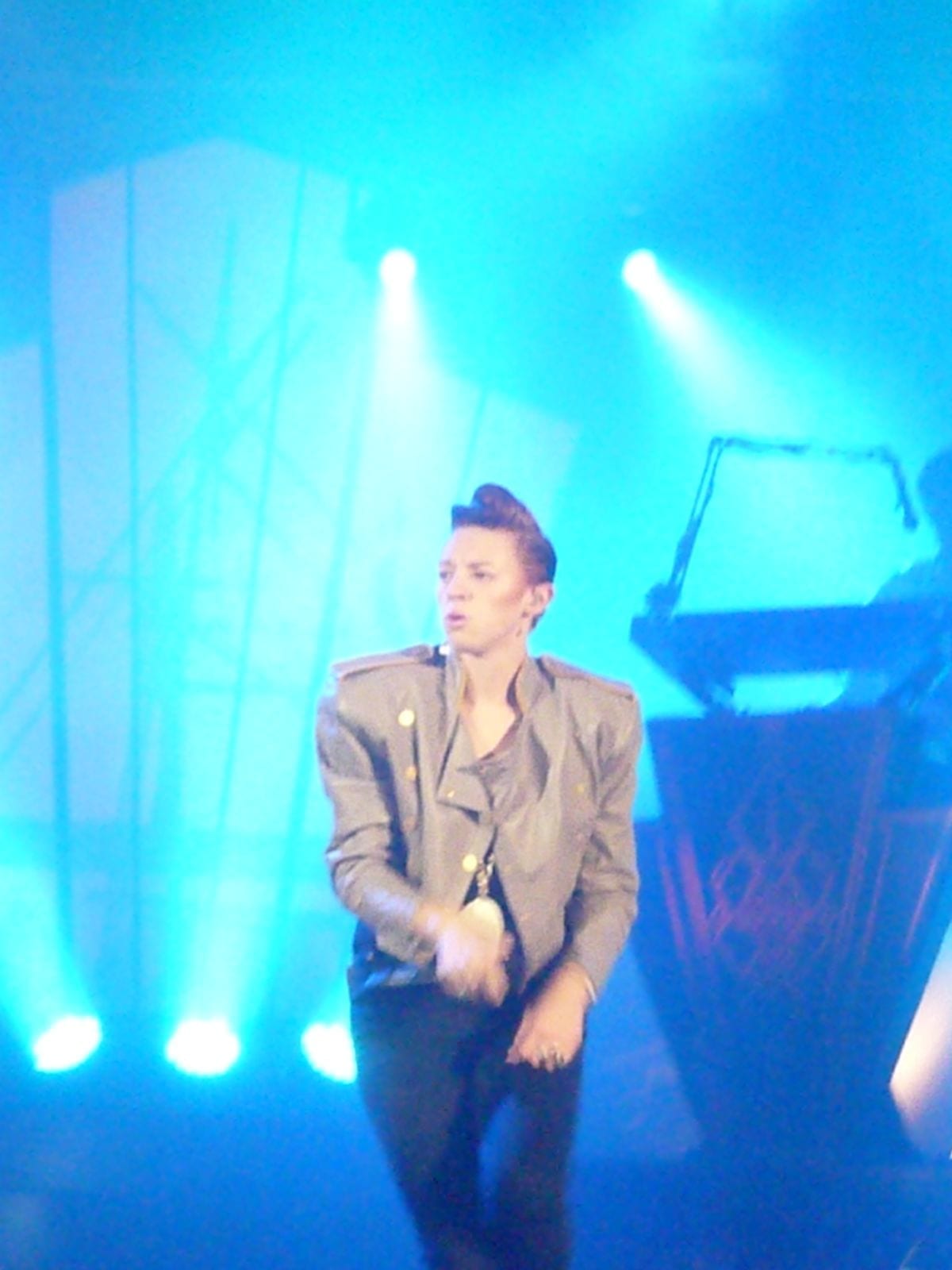 La Roux performs on stage in a gray military-style jacket, singing into a microphone with a pompadour hairstyle, under blue stage lights.