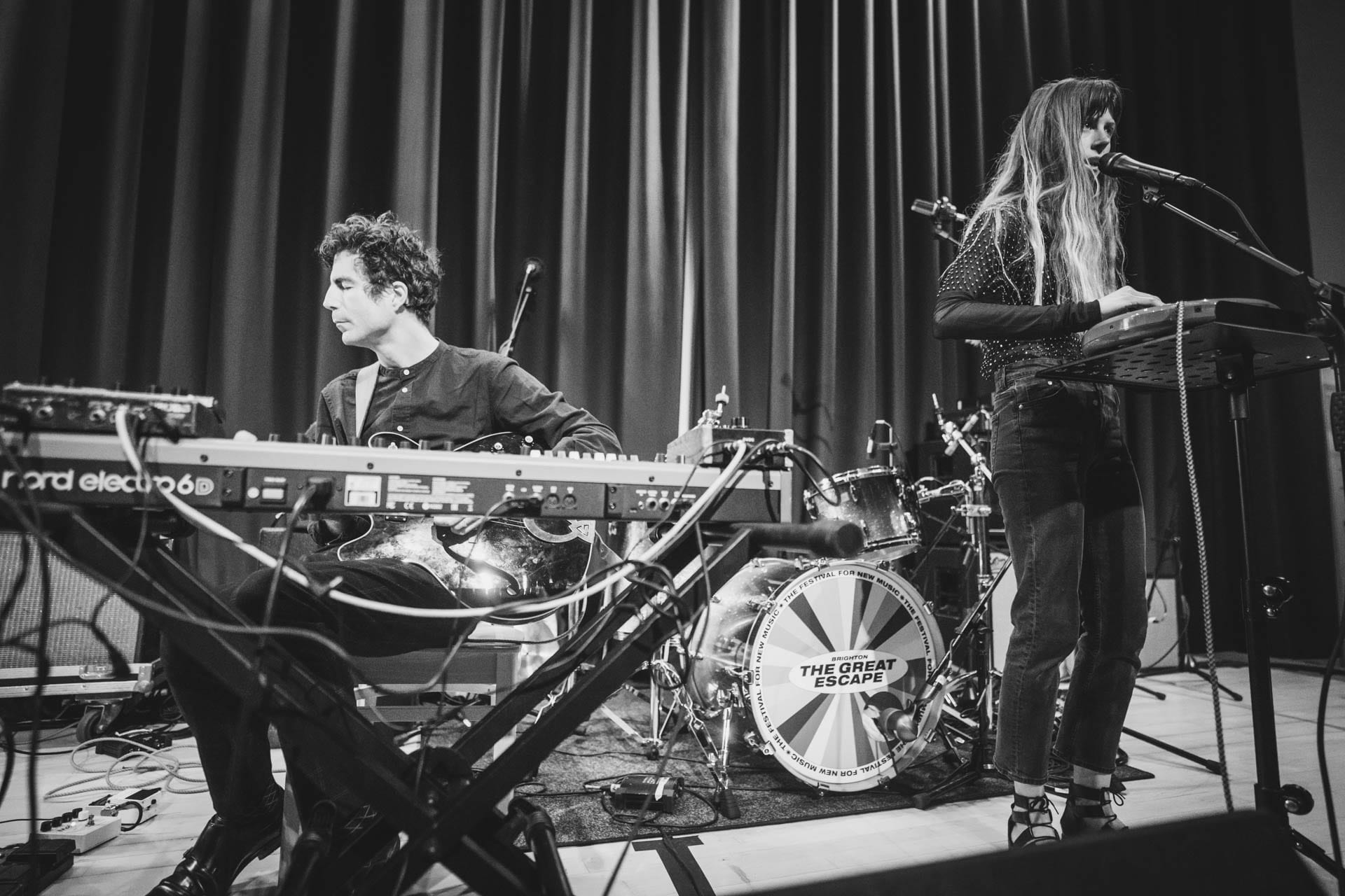 Lael Neale sings into a microphone while playing a small keyboard on stage, in a black and white photo, with a bandmate on synthesizer beside her.