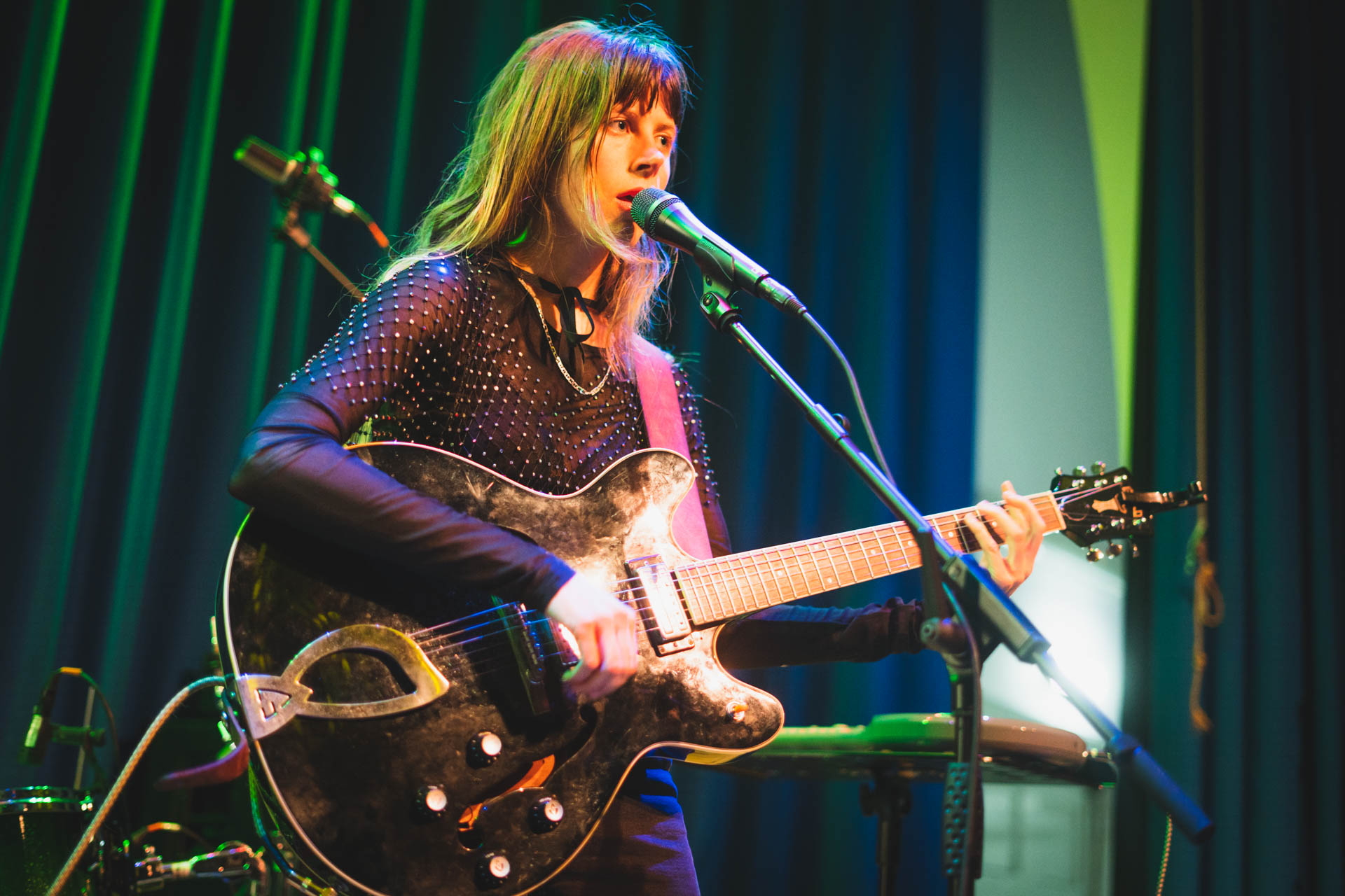 A singer with long fringed hair in a studded top plays a battered hollow-body electric guitar and sings into a mic stand under green and pink light