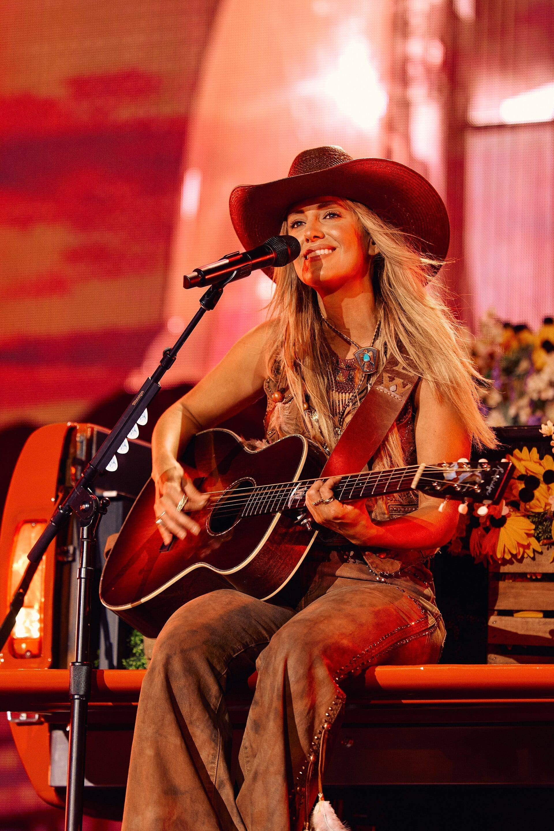 Lainey Wilson smiles while playing an acoustic guitar and singing into a microphone, wearing a wide-brimmed hat and fringed western-style clothing on an orange-lit stage.