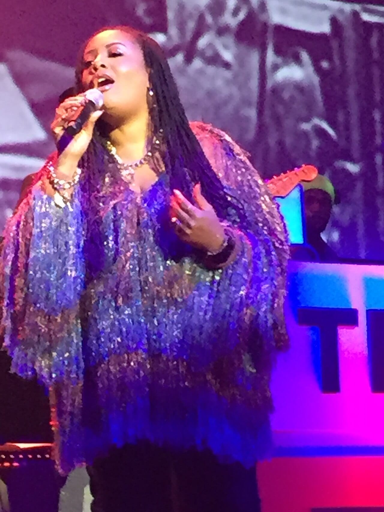 Lalah Hathaway sings into a handheld microphone on stage in a sequinned fringed jacket, lit pink and blue.