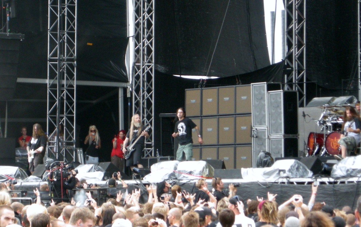 Lamb of God play an outdoor festival stage in daylight, the singer at the centre microphone with the bassist to his left and the drum riser behind, seen over the heads of the crowd.