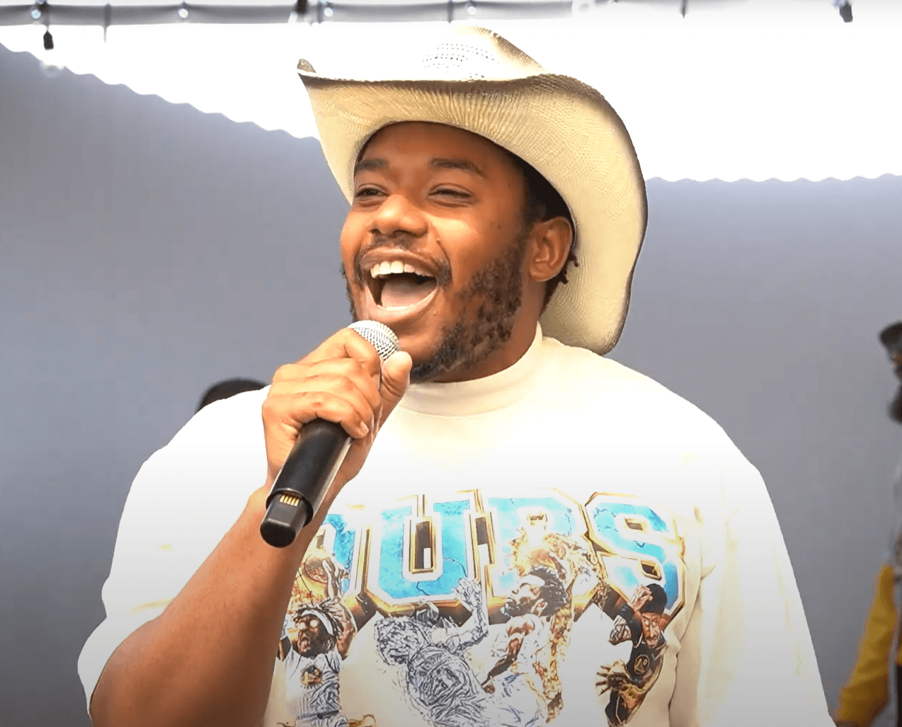 LaRussell laughs while speaking into a handheld microphone, wearing a cream cowboy hat and a white Golden State Warriors graphic sweatshirt.