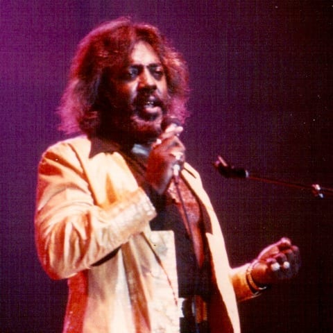 Latimore sings into a handheld microphone in a pale jacket and scarf under warm purple stage light.