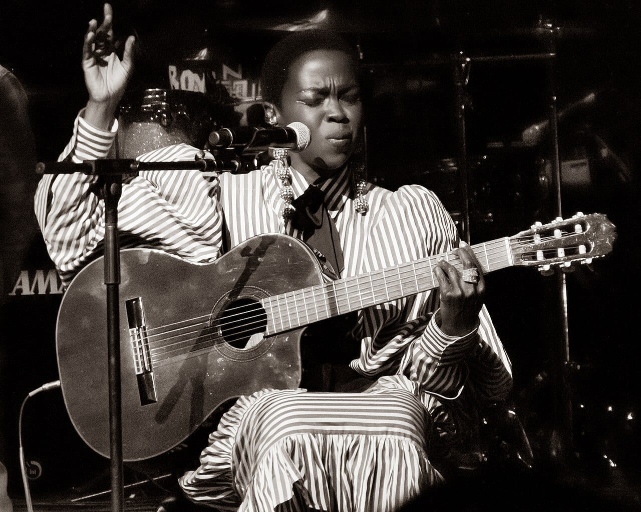 Lauryn Hill sings into a microphone while playing a classical guitar on stage, in a black and white photograph.