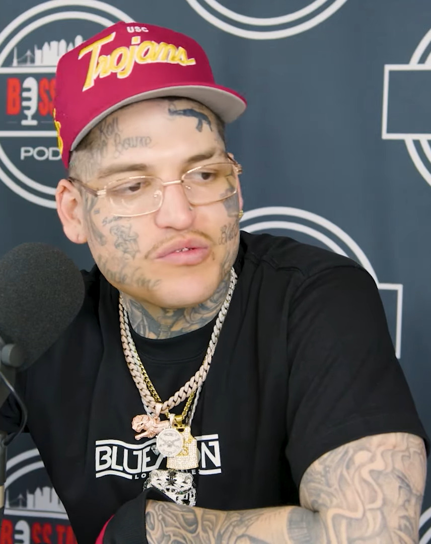 Lefty Gunplay sits behind a podcast microphone in a red USC Trojans cap and gold-rimmed glasses, face and arms heavily tattooed, wearing several diamond chains.
