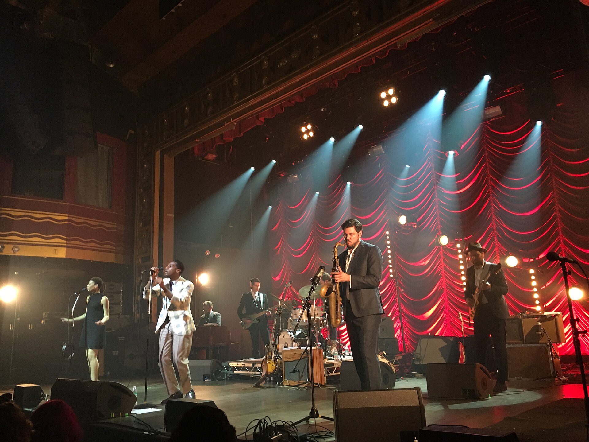 Leon Bridges and his band play a theatre stage lit red, seen from the balcony with the room's ornate arch above them.