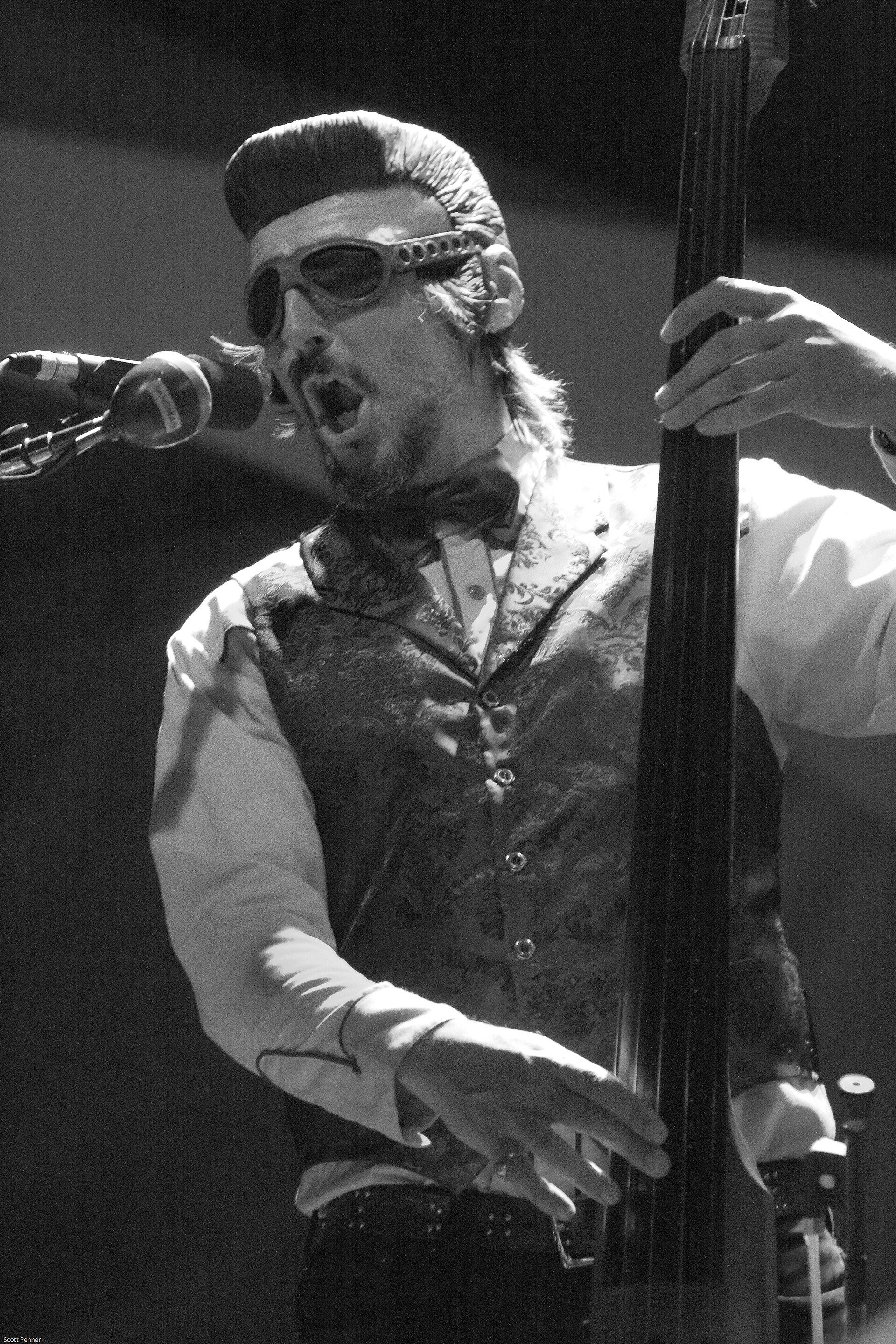 Les Claypool sings into a microphone while playing an upright electric bass, wearing goggles pushed over a quiffed wig, a bow tie and a brocade waistcoat, in black and white.