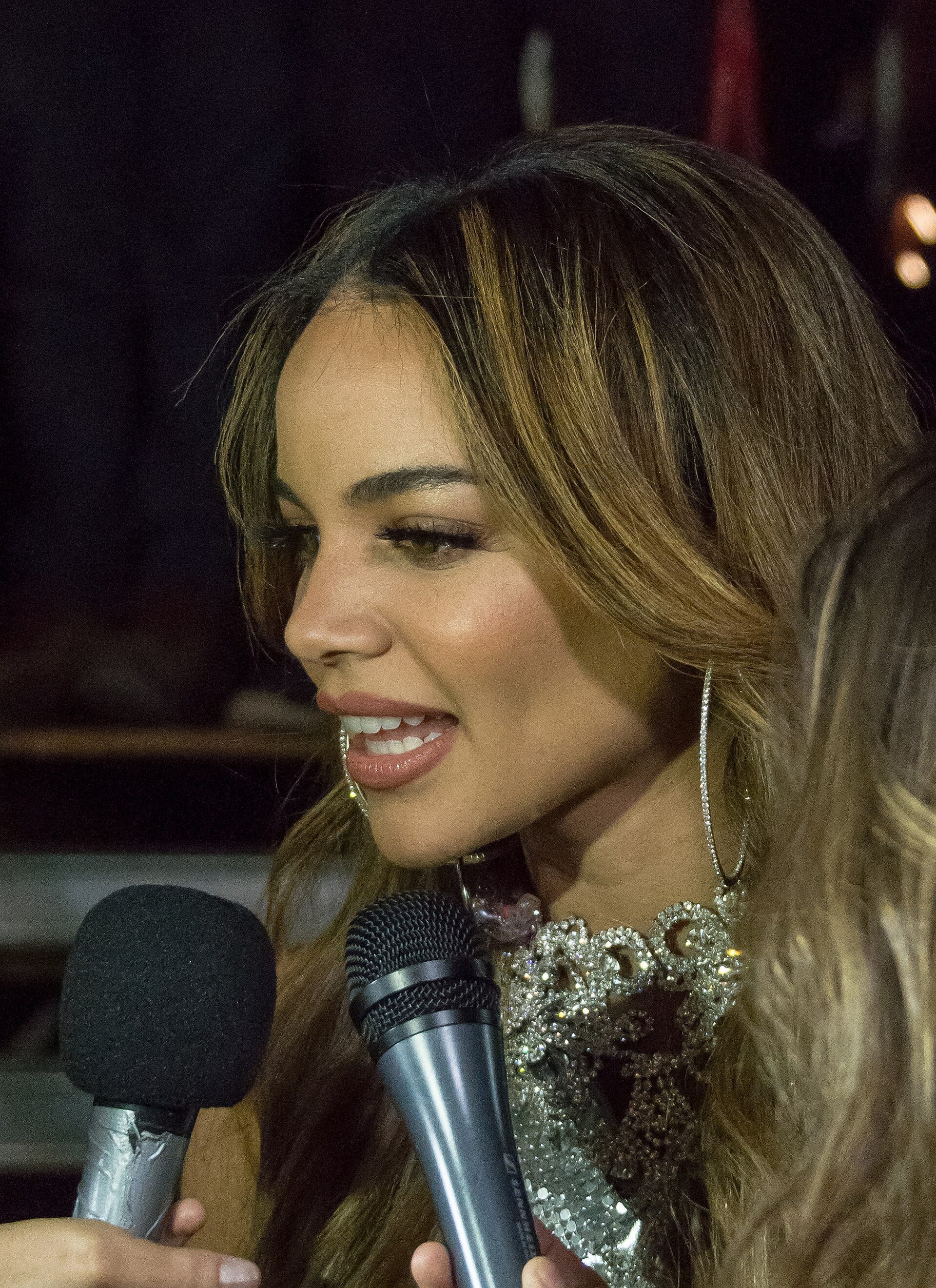 Leslie Grace, in a sparkling silver top and diamond hoop earrings, smiles while speaking into multiple microphones.