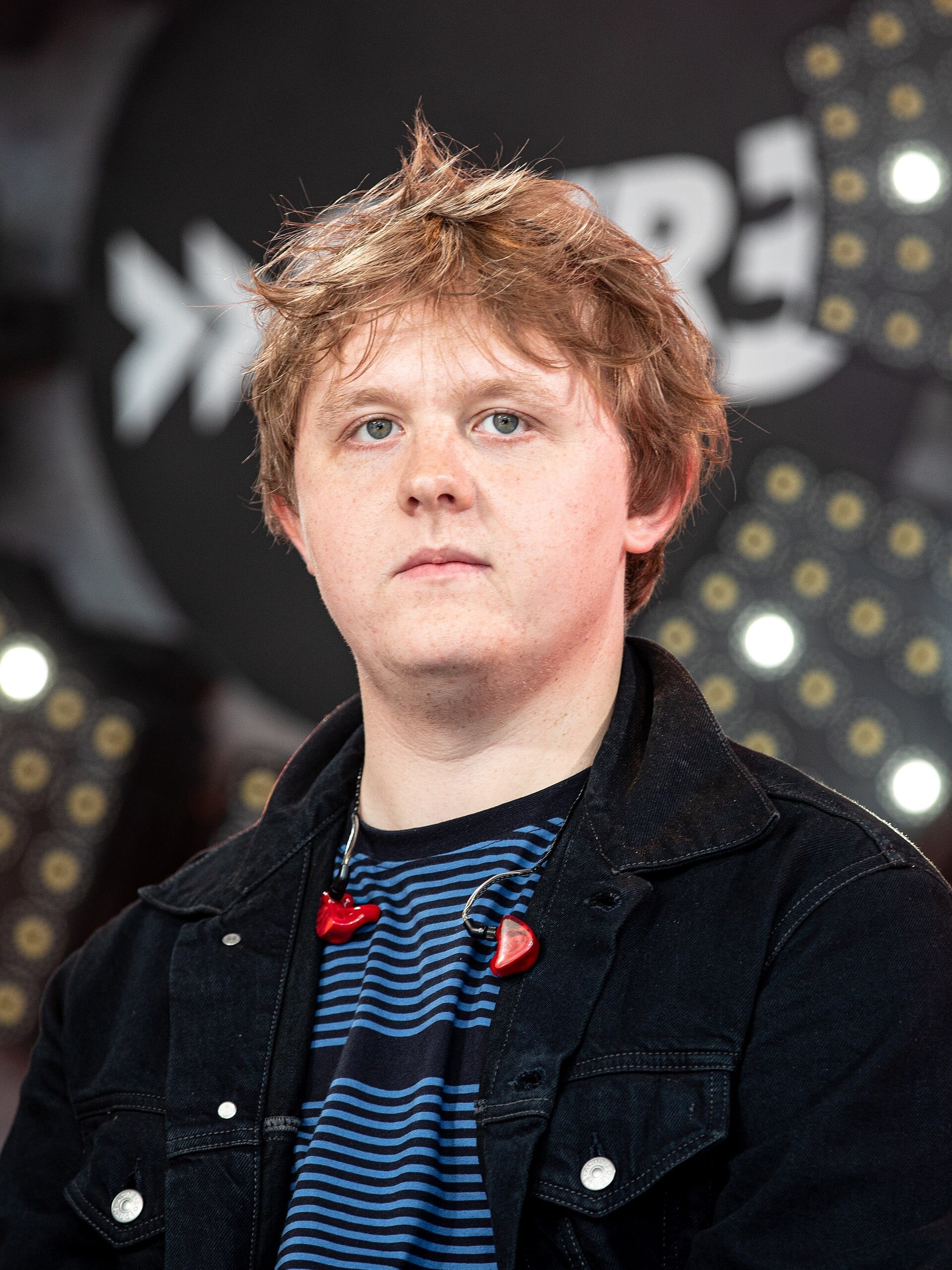 Lewis Capaldi looks at the camera with a neutral expression in a close-up portrait, wearing a black denim jacket over a blue-striped shirt with red in-ear monitors hanging around his neck.