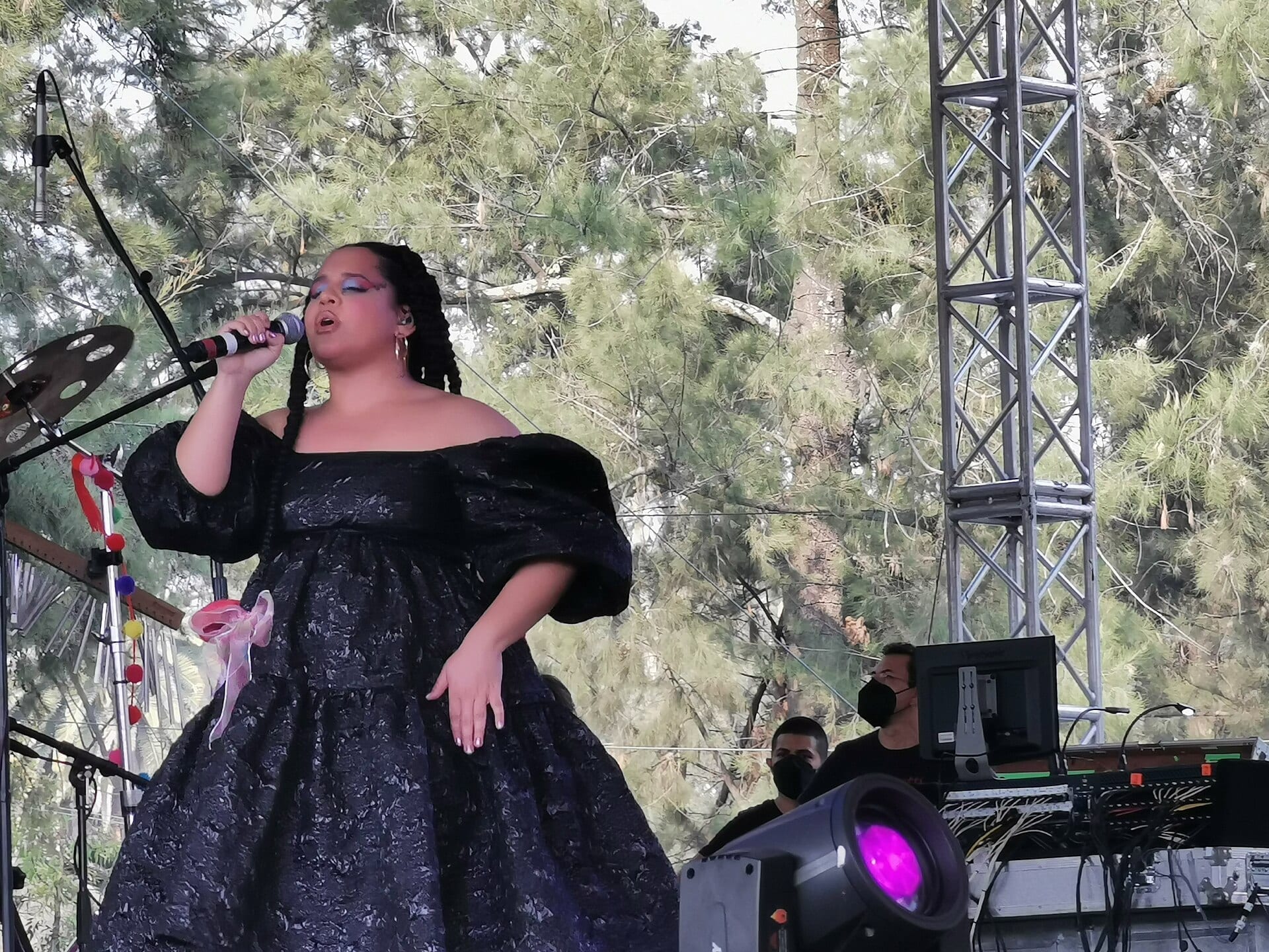 Lido Pimienta, in an elaborate black off-shoulder gown and braided hair, sings into a microphone on an outdoor festival stage.