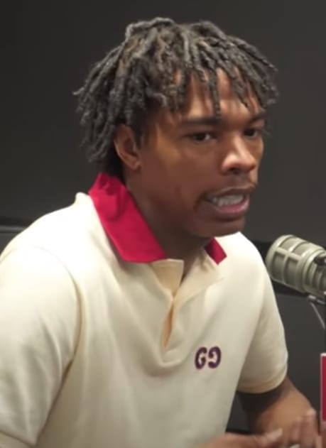 Close-up of Lil Baby speaking into a microphone during an interview, wearing a cream and red polo shirt with dreadlocked hair.