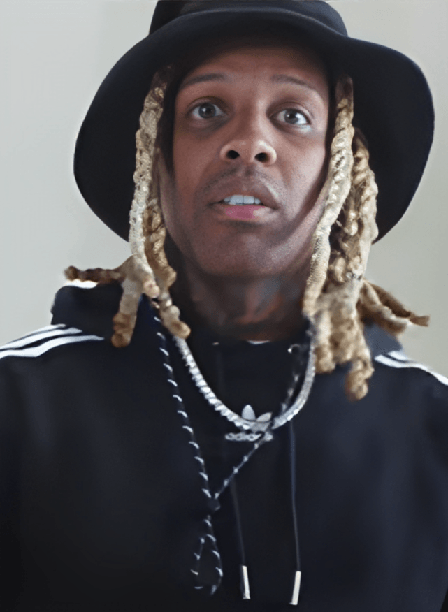 Lil Durk, a Black man with long light-blond dreadlocks, wearing a black wide-brimmed hat, a black hoodie, and a silver chain necklace, looking toward the camera.
