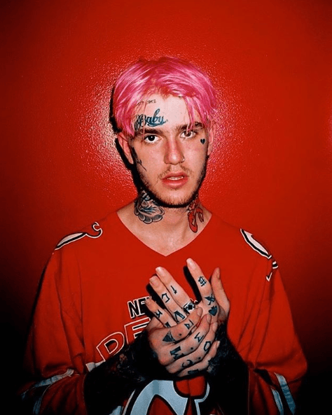 Lil Peep, with pink hair and face tattoos including a heart under one eye, poses against a red background wearing a red hockey jersey and holding his tattooed hands together.