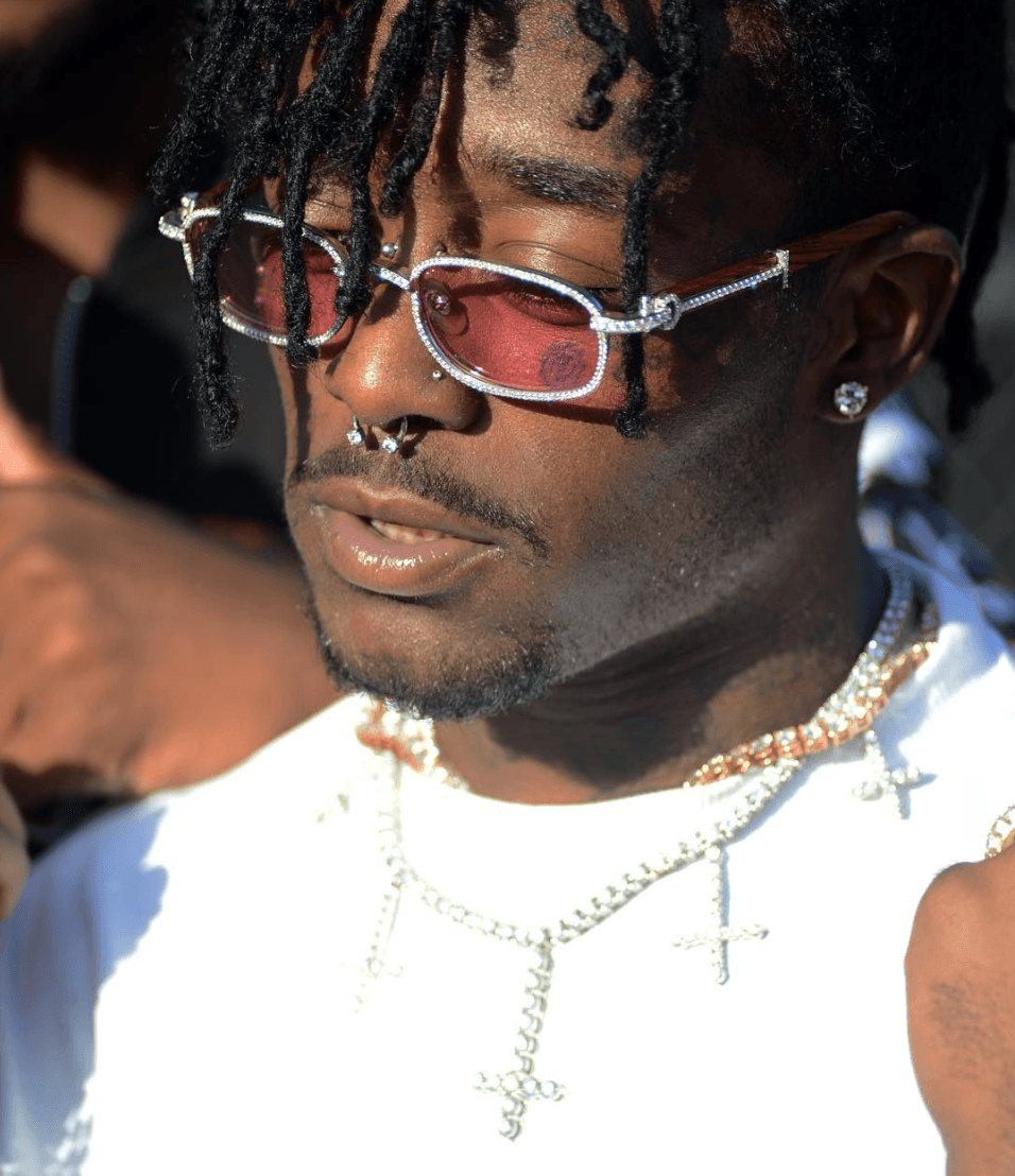 Lil Uzi Vert in close-up outdoor sunlight, wearing pink-tinted rectangular sunglasses with rhinestone trim, dreadlocks, a septum piercing, and layered silver chains over a white tank top.