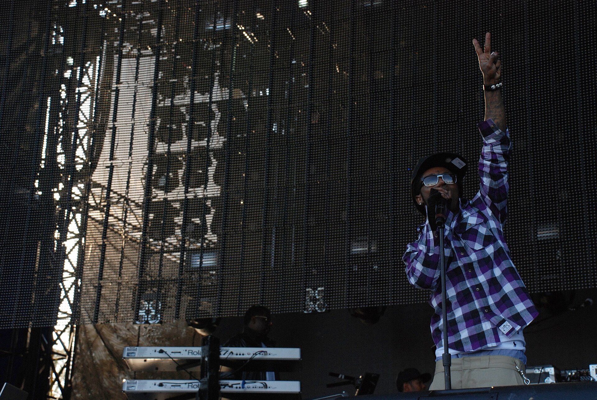 Lil Wayne raises one hand above his head at a microphone stand, in a purple and white checked shirt, dark cap and sunglasses, in front of a large stage screen.