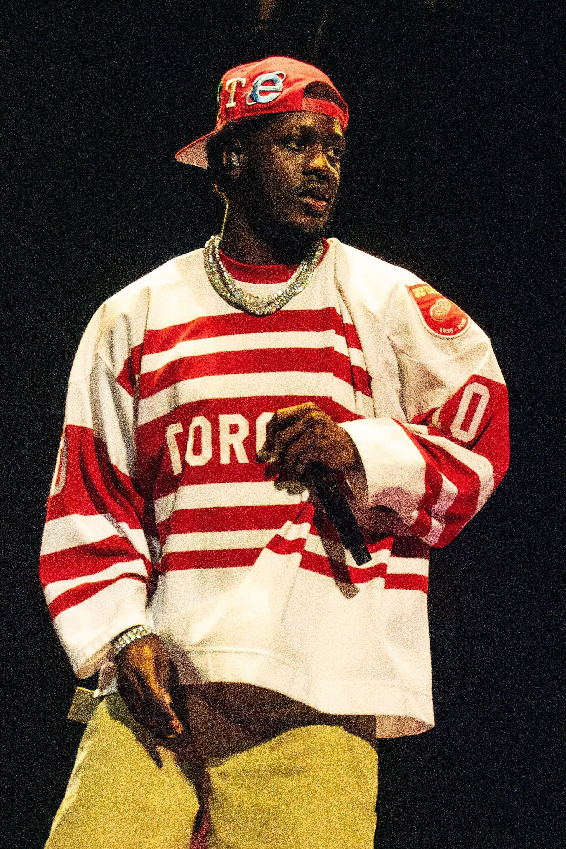 Lil Yachty holds a microphone in a red and white striped Toronto hockey jersey and a backwards red cap, with heavy chains at his neck and wrist.