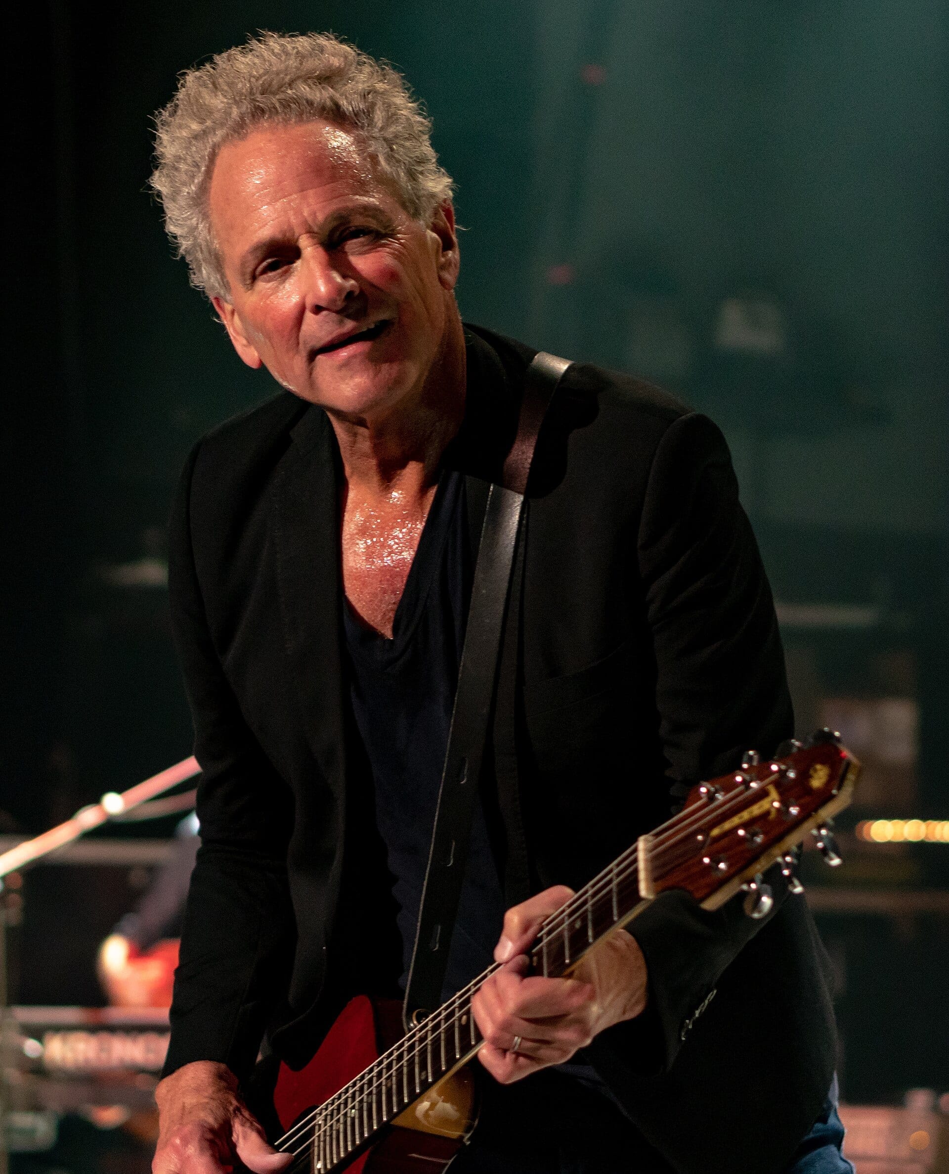Lindsey Buckingham performs on stage in a black jacket, smiling while playing an electric guitar under stage lighting.