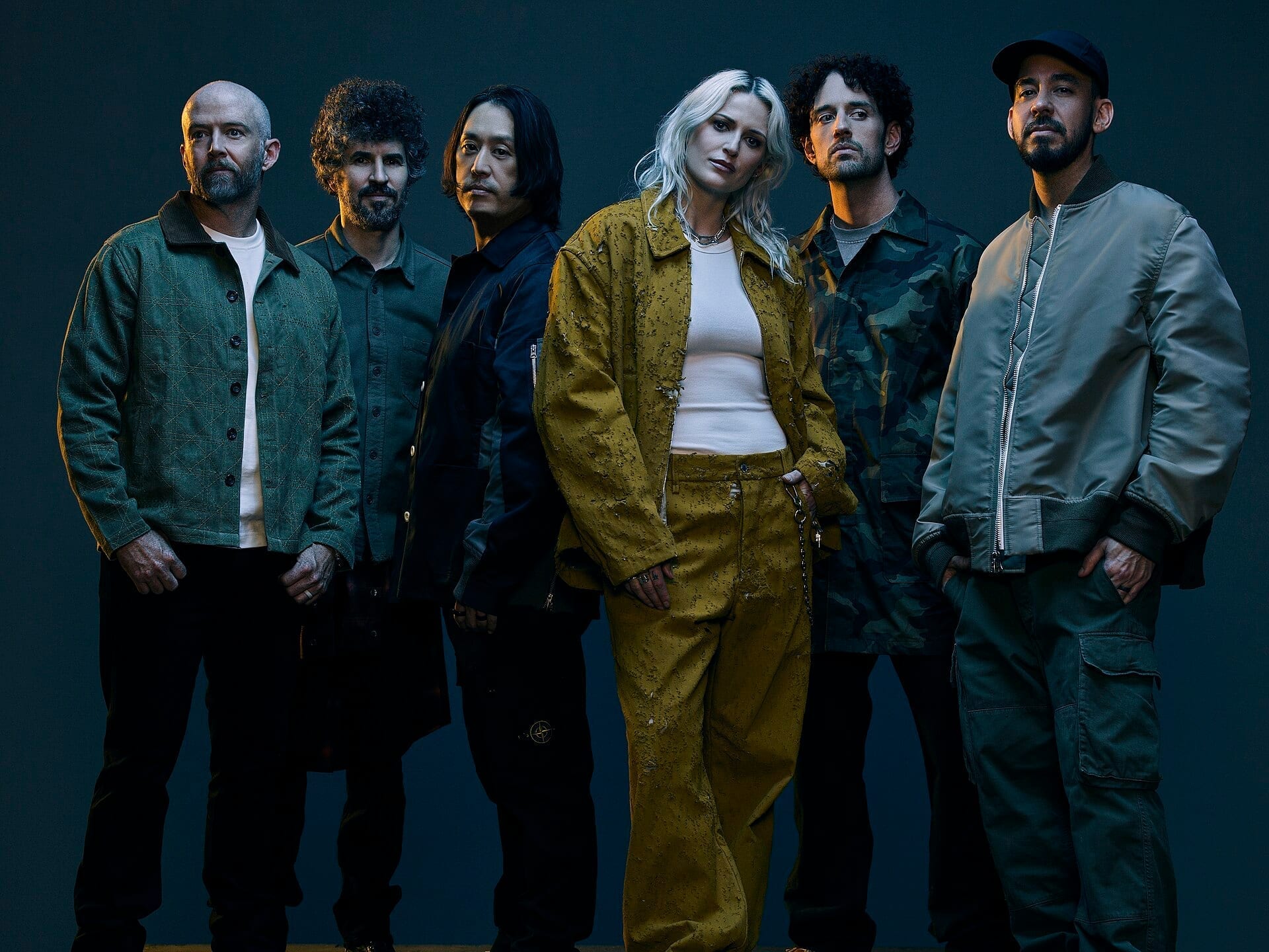 The six members of Linkin Park stand together for a group studio portrait against a dark grey background, dressed in muted green, camouflage, and navy jackets, with a blonde woman in a mustard-yellow jacket standing at the center.