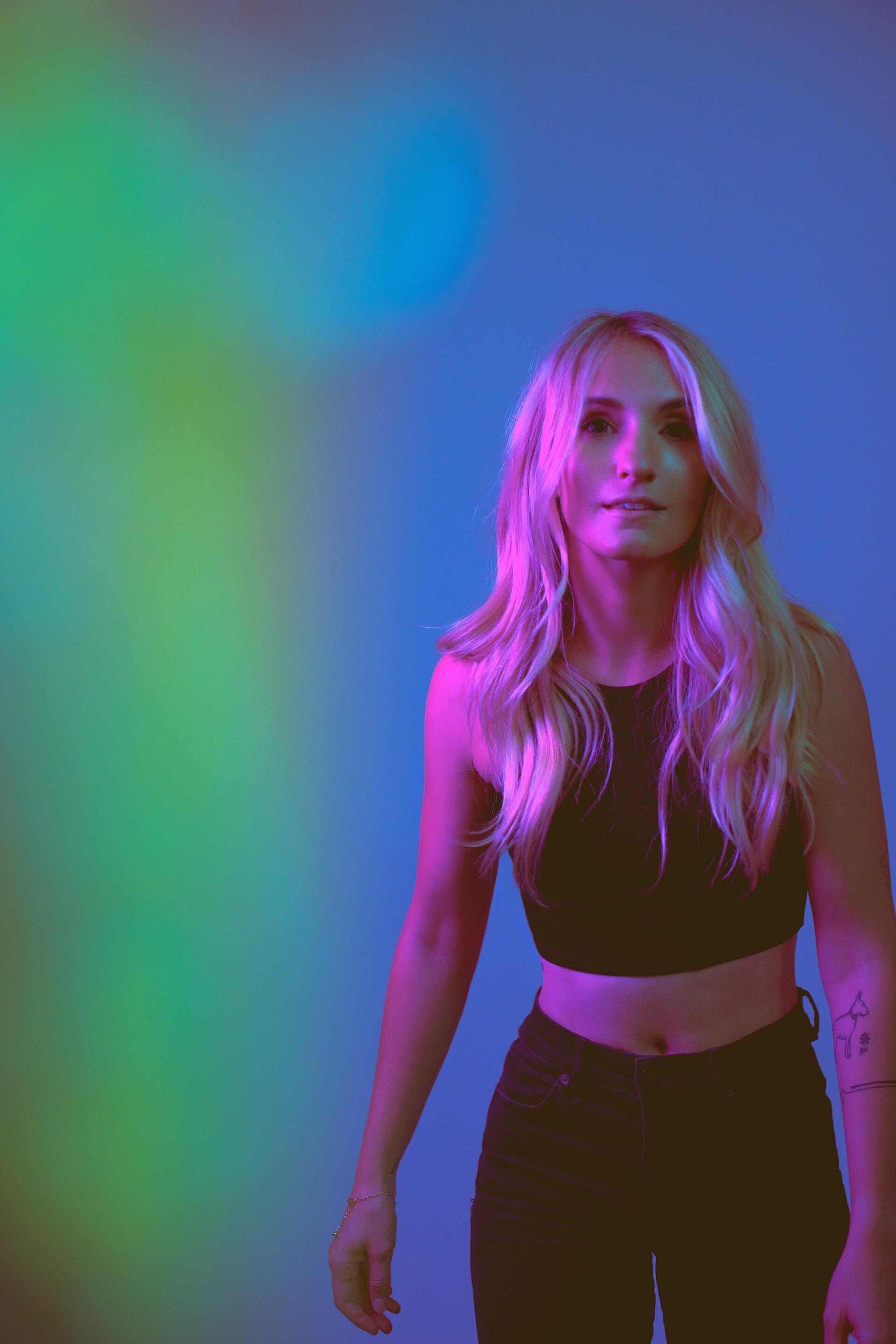 Linney, a blonde woman in a black crop top and jeans, stands under blue and green stage lighting.