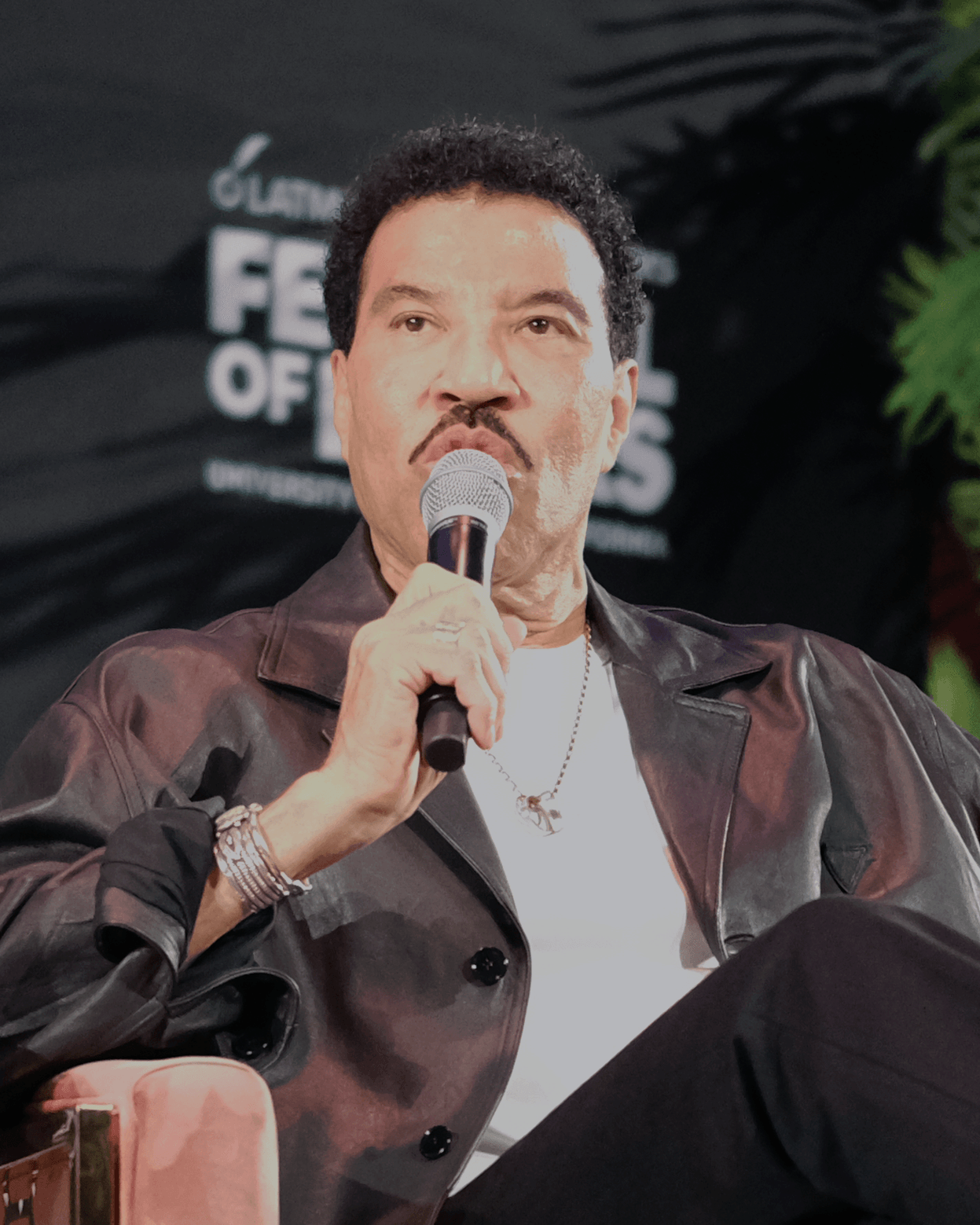 Lionel Richie in a dark jacket and open collar, speaking with a microphone in front of him.
