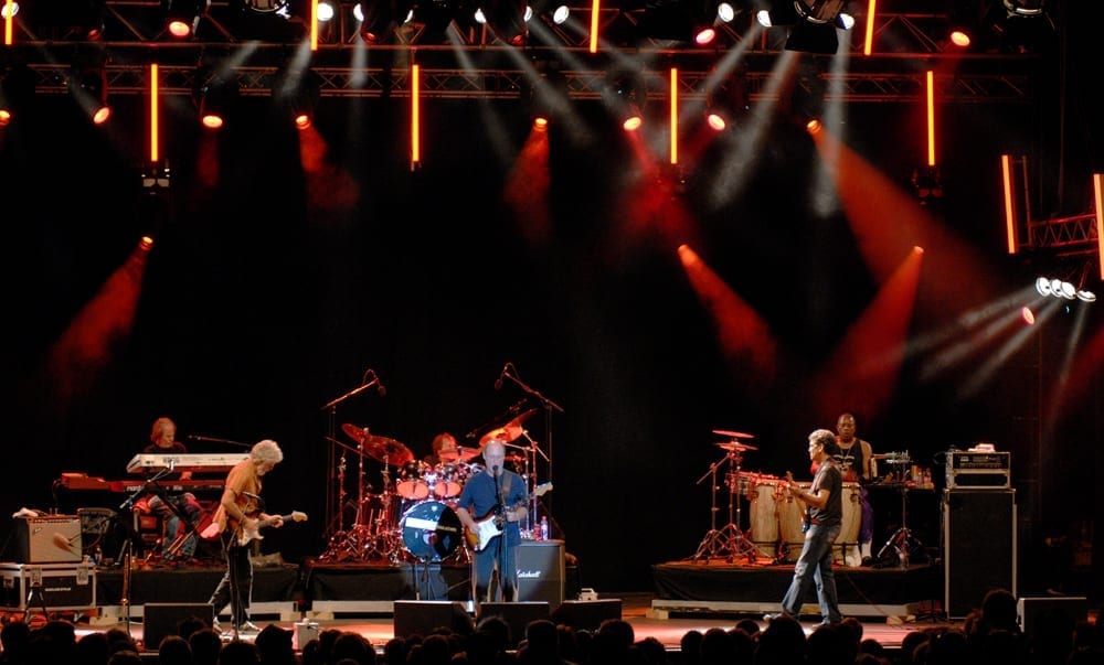 Little Feat performs across a wide stage under fanned red and white spotlights, with a keyboardist, guitarists, drummer and percussionists spread out.