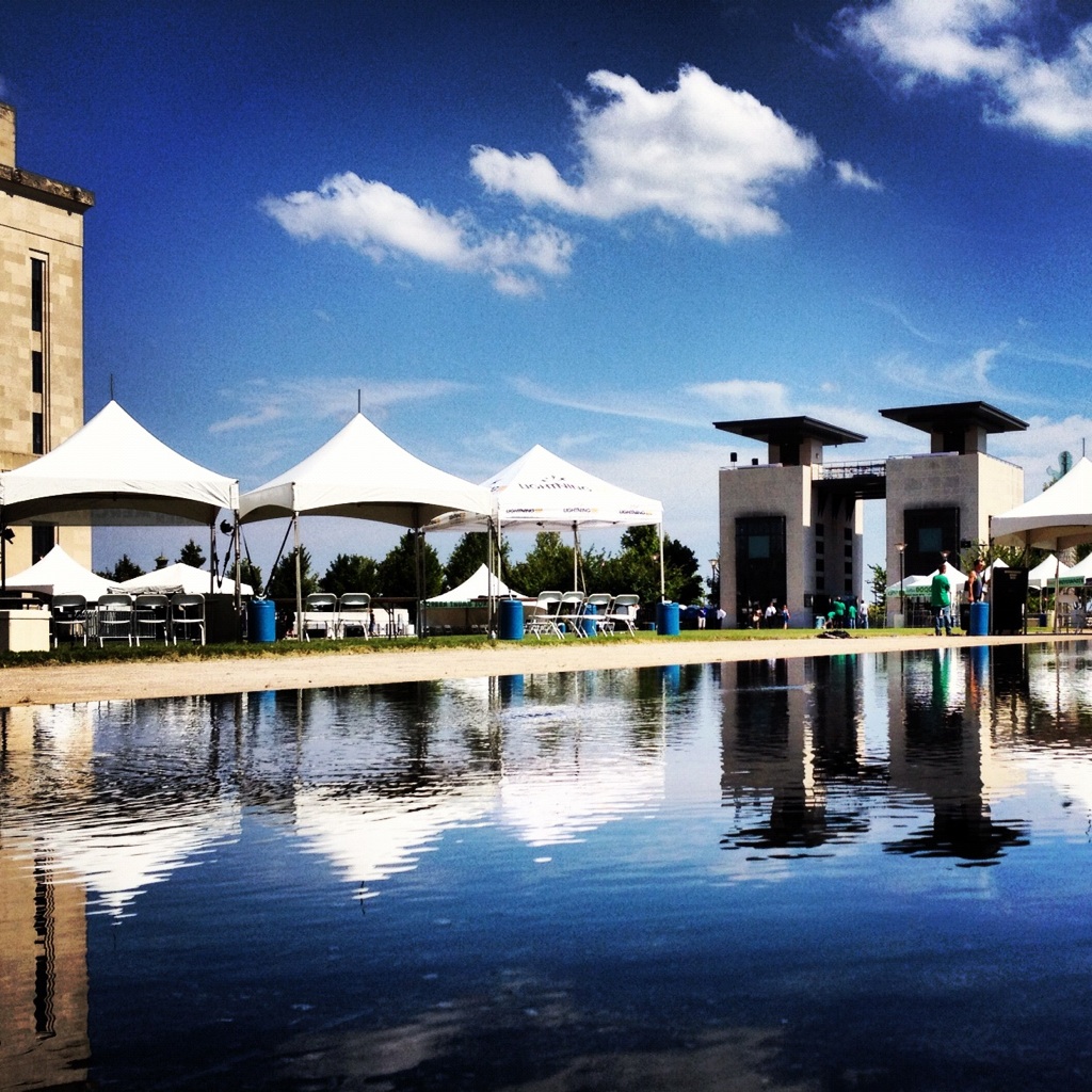 White event tents and two dark stone towers stand on a lawn under a blue sky, mirrored in a shallow reflecting pool that fills the foreground.