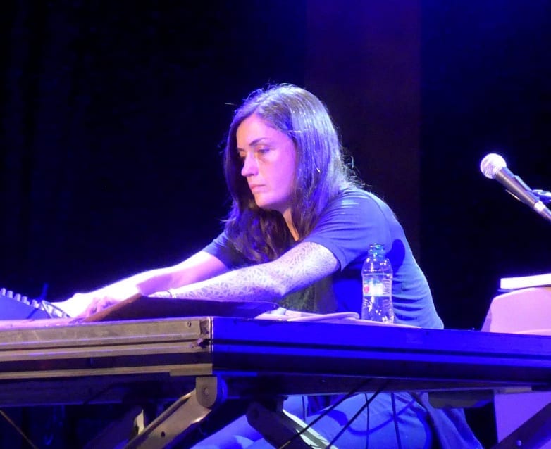 Liz Harris, lit in blue stage light, playing a keyboard with her hair falling over her face.