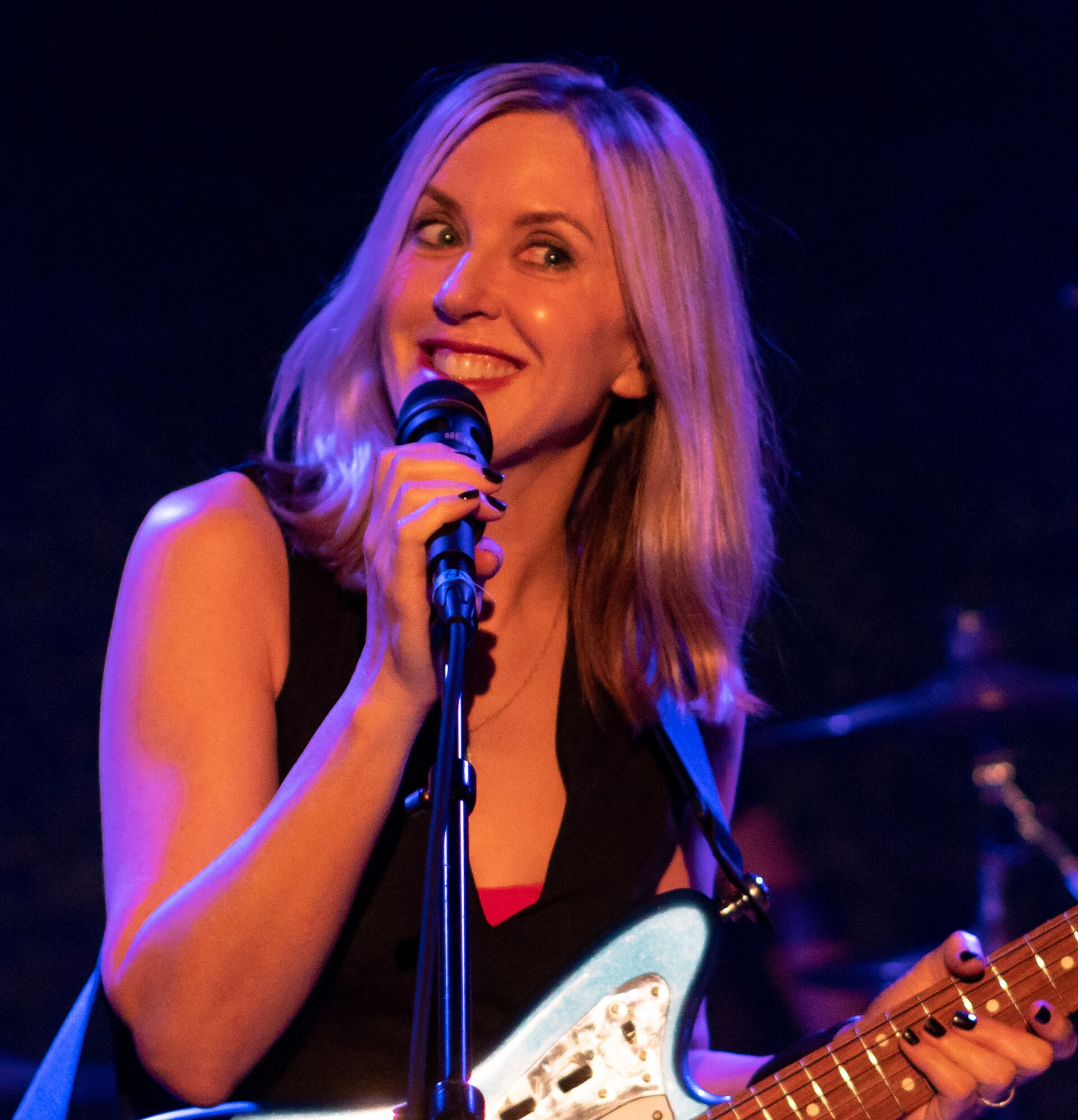 Liz Phair sings into a microphone on a stand with her eyes half closed, an electric guitar slung over her black sleeveless top.