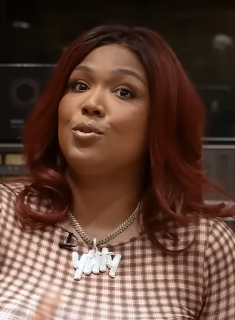 Lizzo talks mid-sentence in a studio, auburn hair past her shoulders, wearing a checked top and a chain necklace with a nameplate pendant.