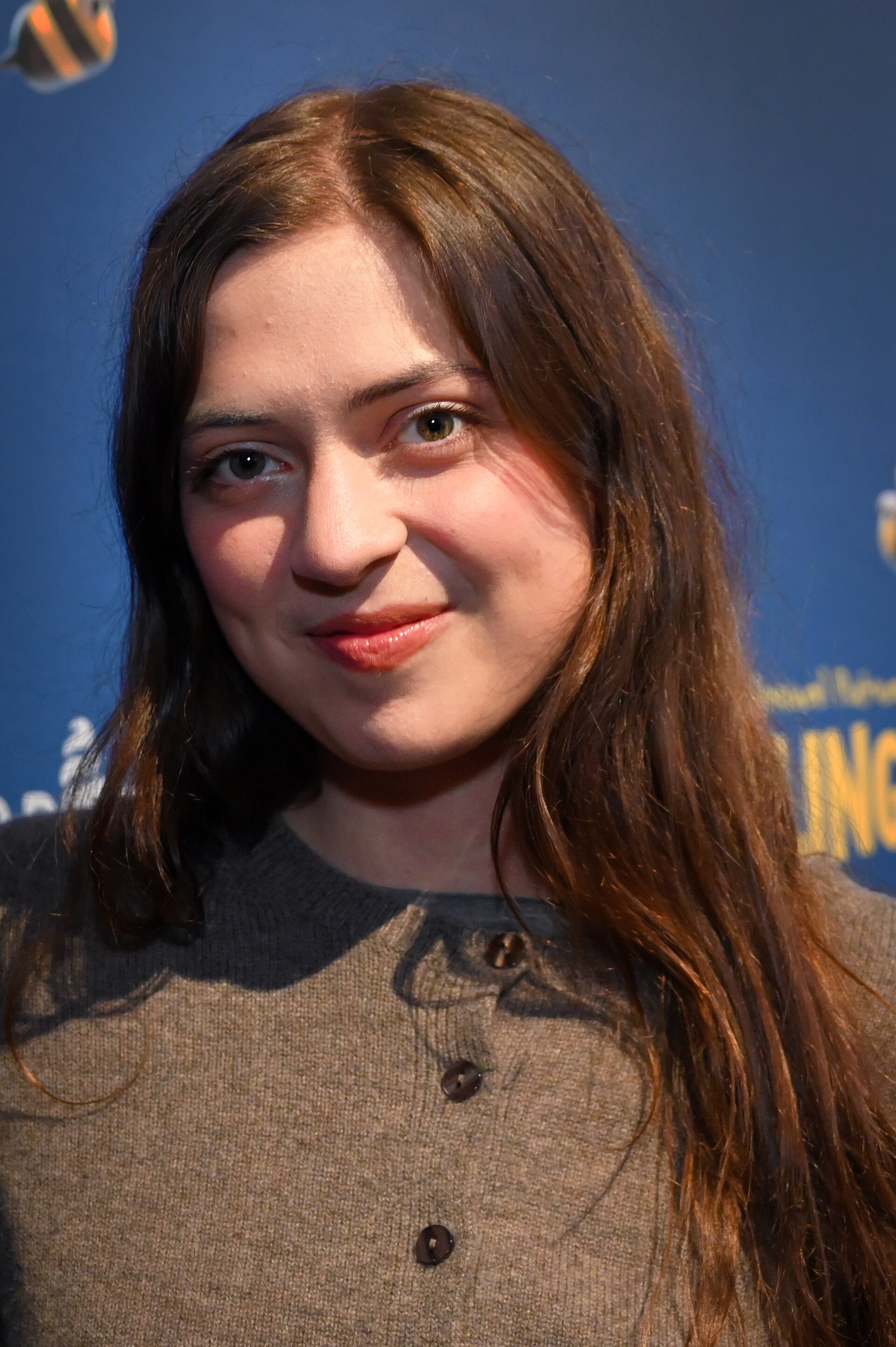 A young woman with long brown hair in a grey cardigan smiles slightly at the camera in front of a blue step-and-repeat backdrop