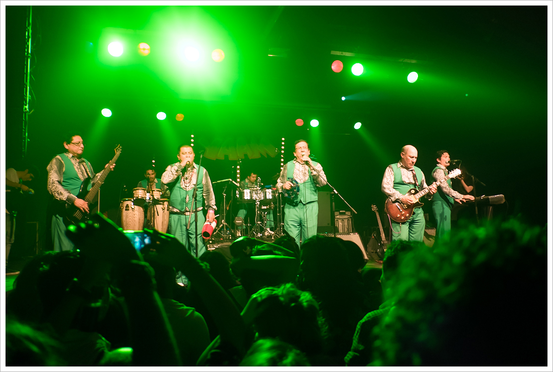 Los Mirlos play a club stage under green light in matching mint waistcoats and trousers, two singers at microphones with a bassist, guitarist, congas and timbales around them, over the heads of the crowd.