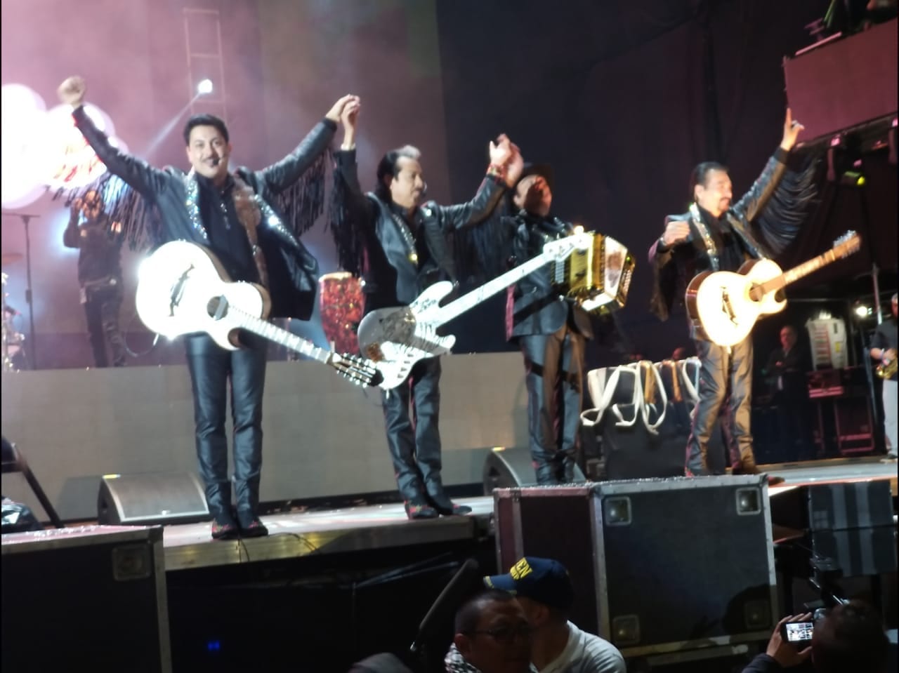 Los Tigres del Norte, in matching black stage jackets, raise their arms on stage with rhinestone-studded guitars and an accordion.