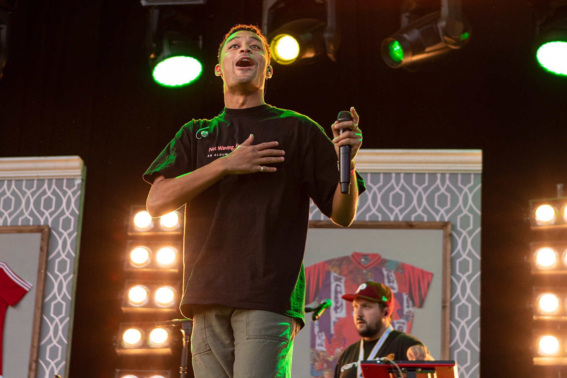 Loyle Carner sings with his head tipped back and one hand flat on his chest, holding a microphone, lit green and amber, with a DJ working behind him.