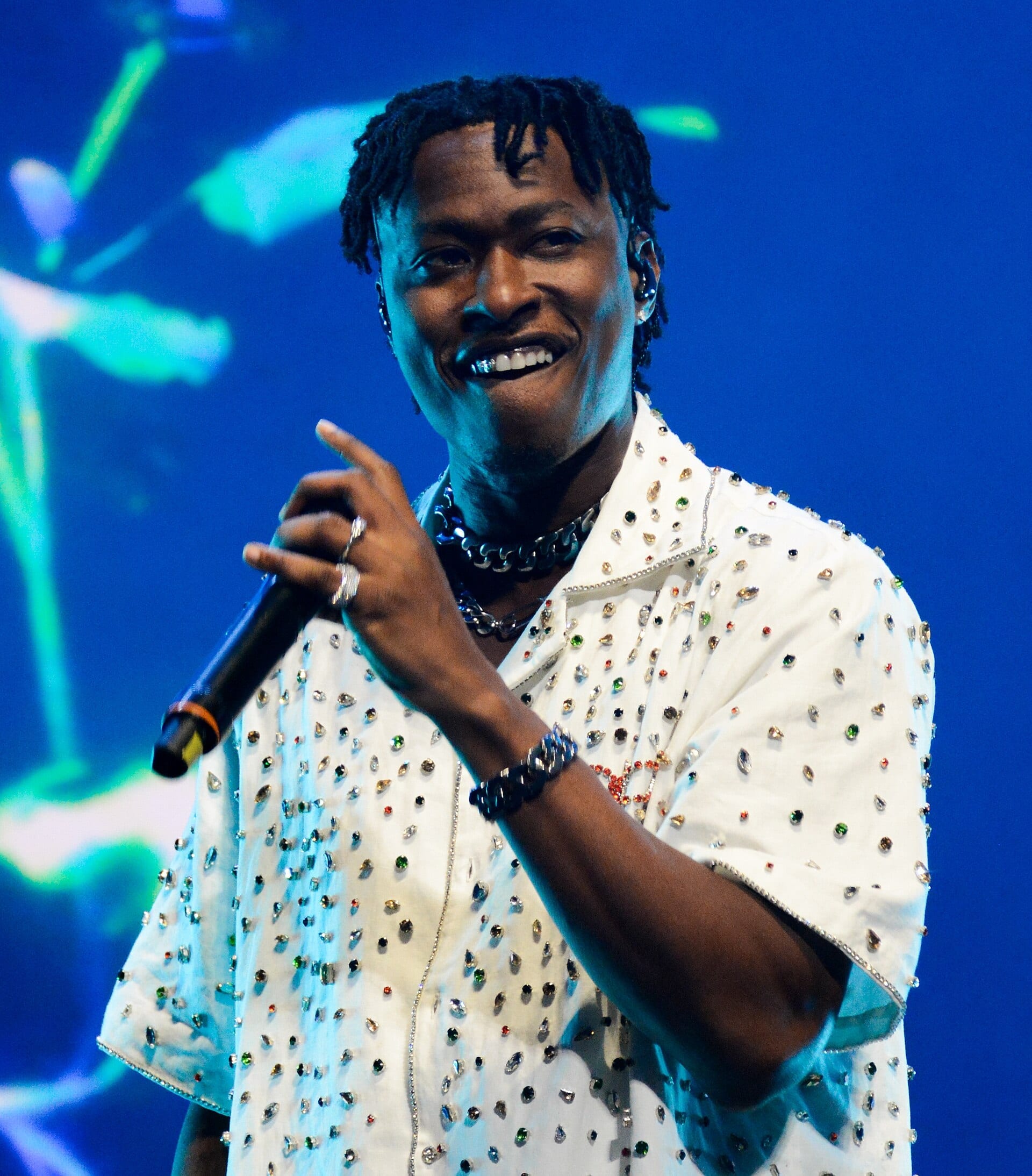 Lucky Daye grins and points with the hand holding his microphone, in a white shirt covered in coloured rhinestones.
