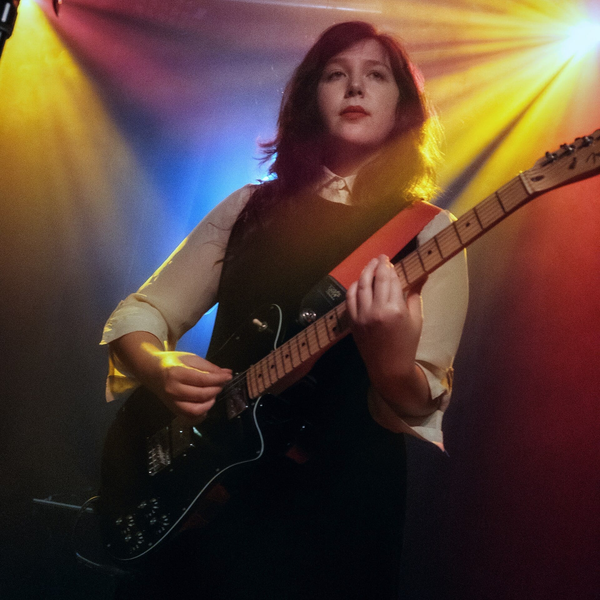 Lucy Dacus plays a red-and-black electric guitar on a dimly lit stage, purple and gold lighting behind her.