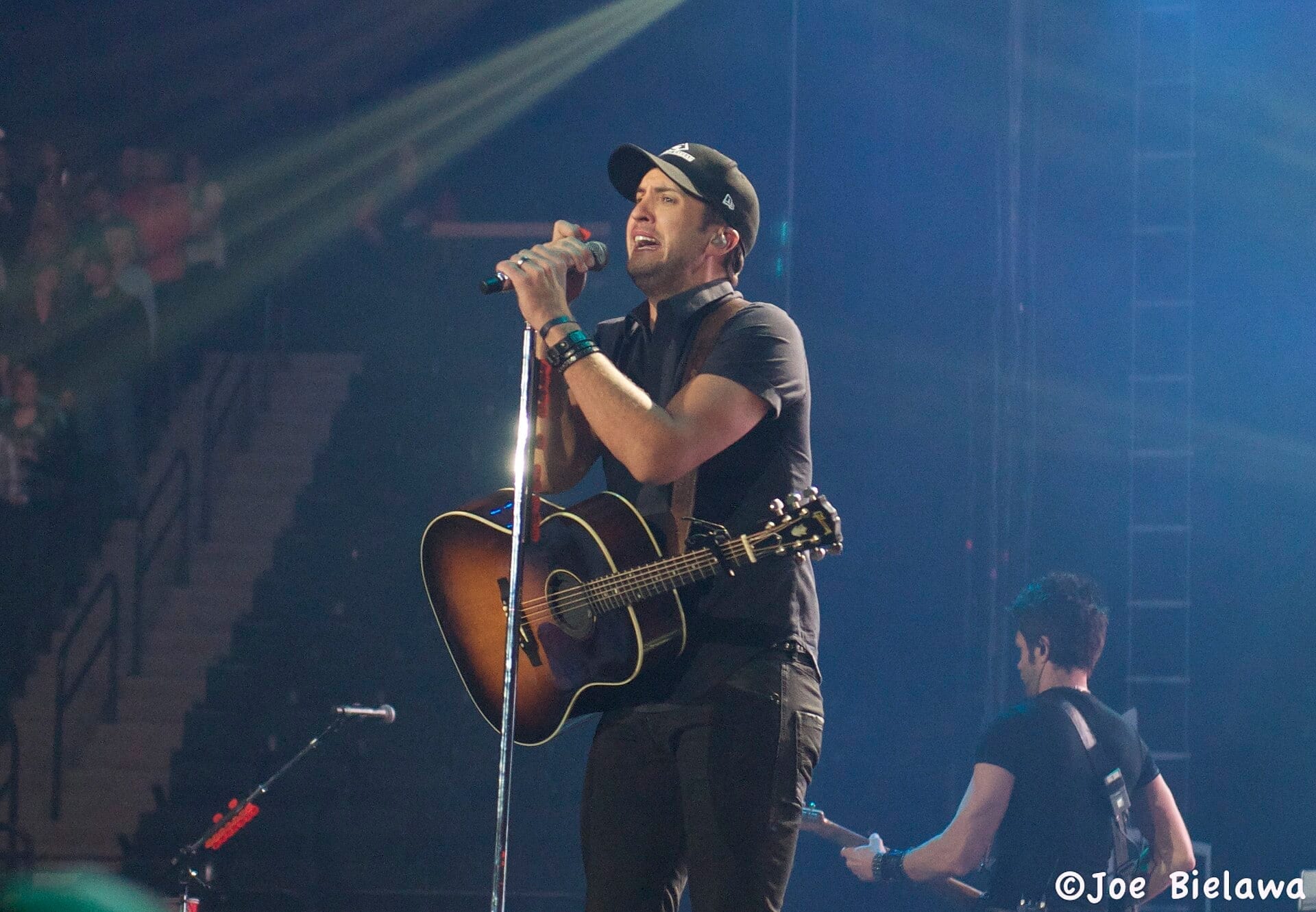 Luke Bryan sings into a microphone while playing an acoustic guitar on stage, wearing a ball cap and dark button-down shirt.