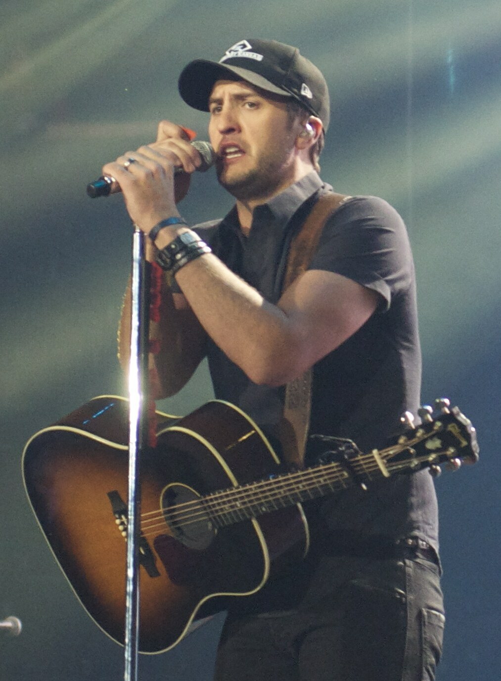 A singer in a baseball cap and grey shirt sings into a microphone on a stand with an acoustic guitar slung across him