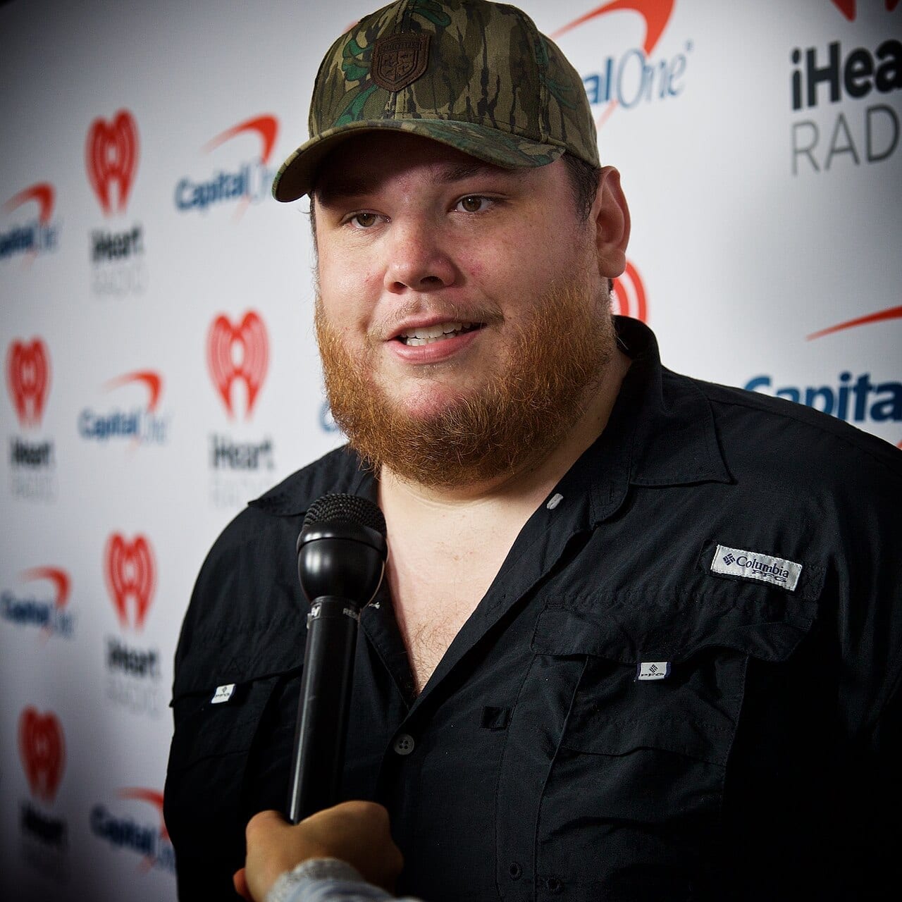 Luke Combs speaks into a handheld microphone in front of a sponsor banner, wearing a camouflage cap.