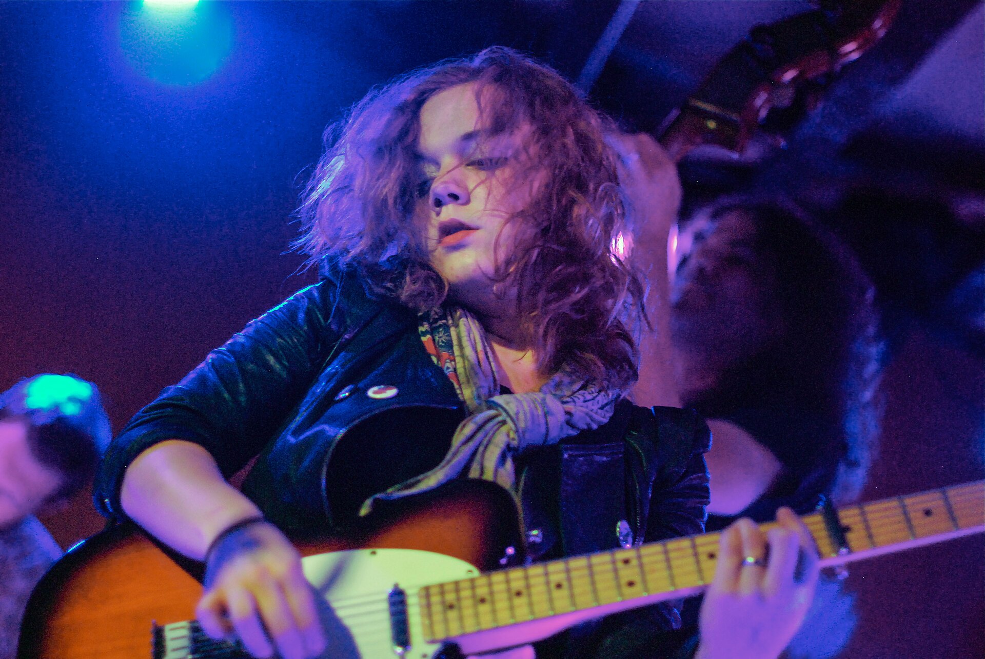 Lydia Loveless playing a Telecaster on a small club stage under blue light, hair falling across her face, a scarf knotted at her neck