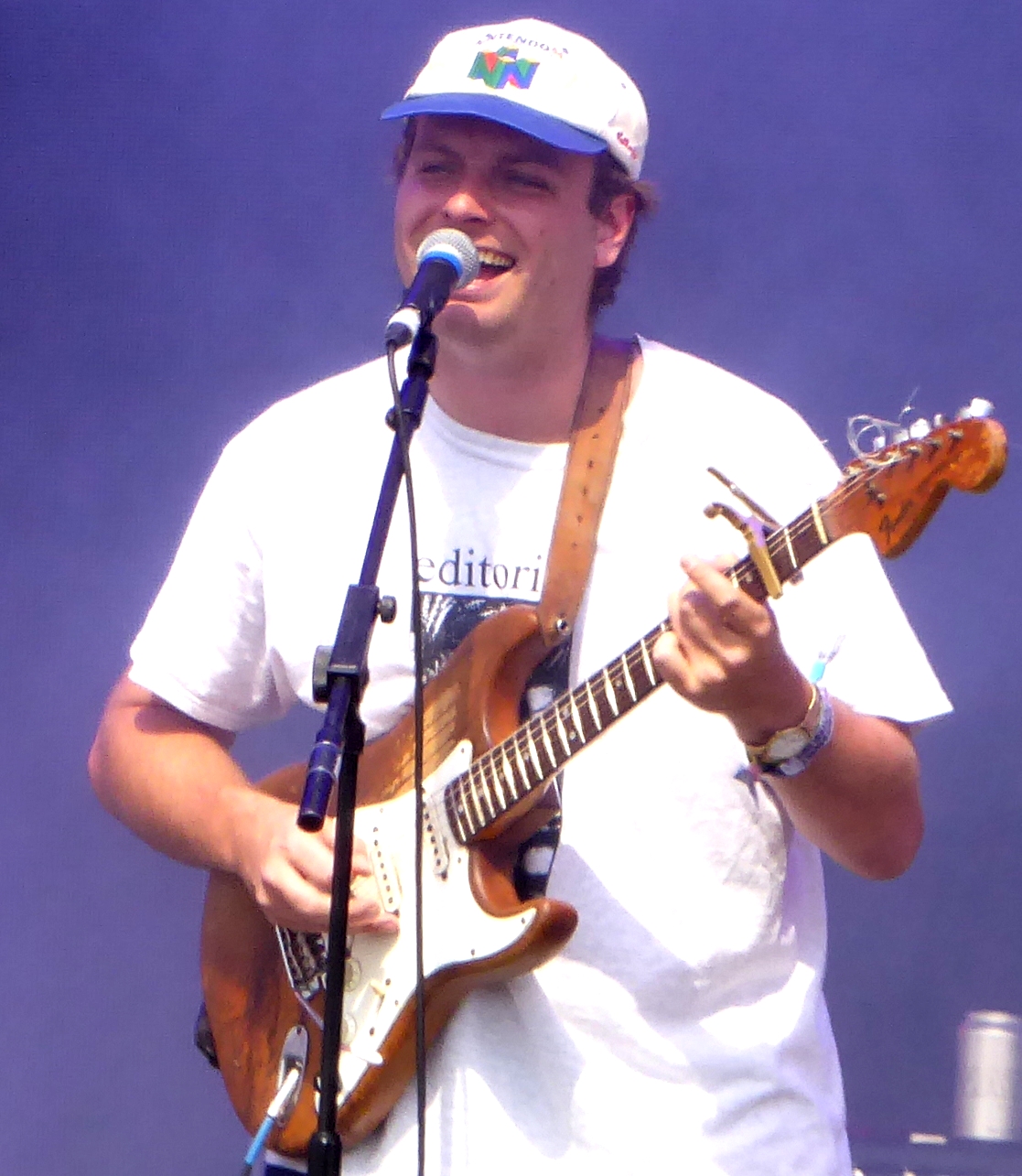 Mac DeMarco grins gap-toothed on an outdoor stage in a baseball cap and a loose shirt, an electric guitar slung across him.