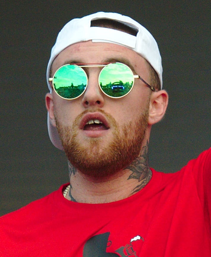 Mac Miller in a backwards white cap and round green mirrored sunglasses, mid-word, in a red T-shirt.