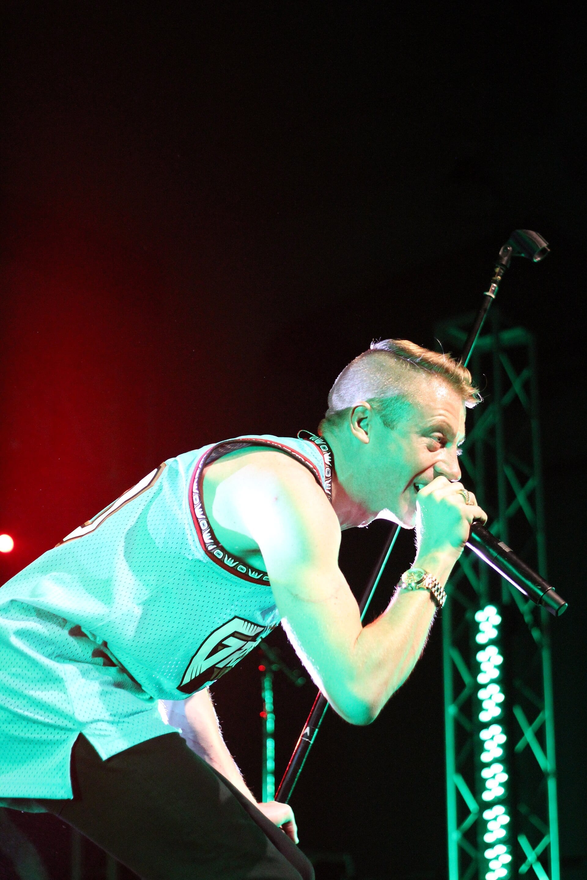Macklemore, with a shaved-side mohawk hairstyle, leans into a microphone mid-performance under green stage lighting, wearing a teal basketball jersey.