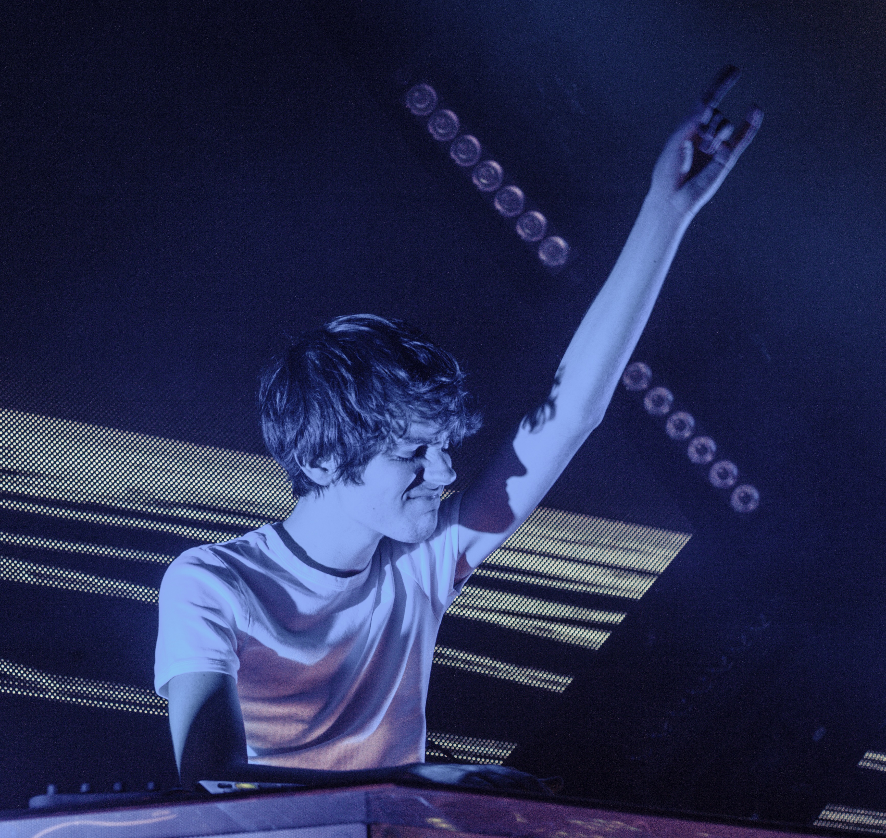 Madeon throws one arm up behind a DJ booth in a white T-shirt, lit blue in front of a screen of horizontal light bars.